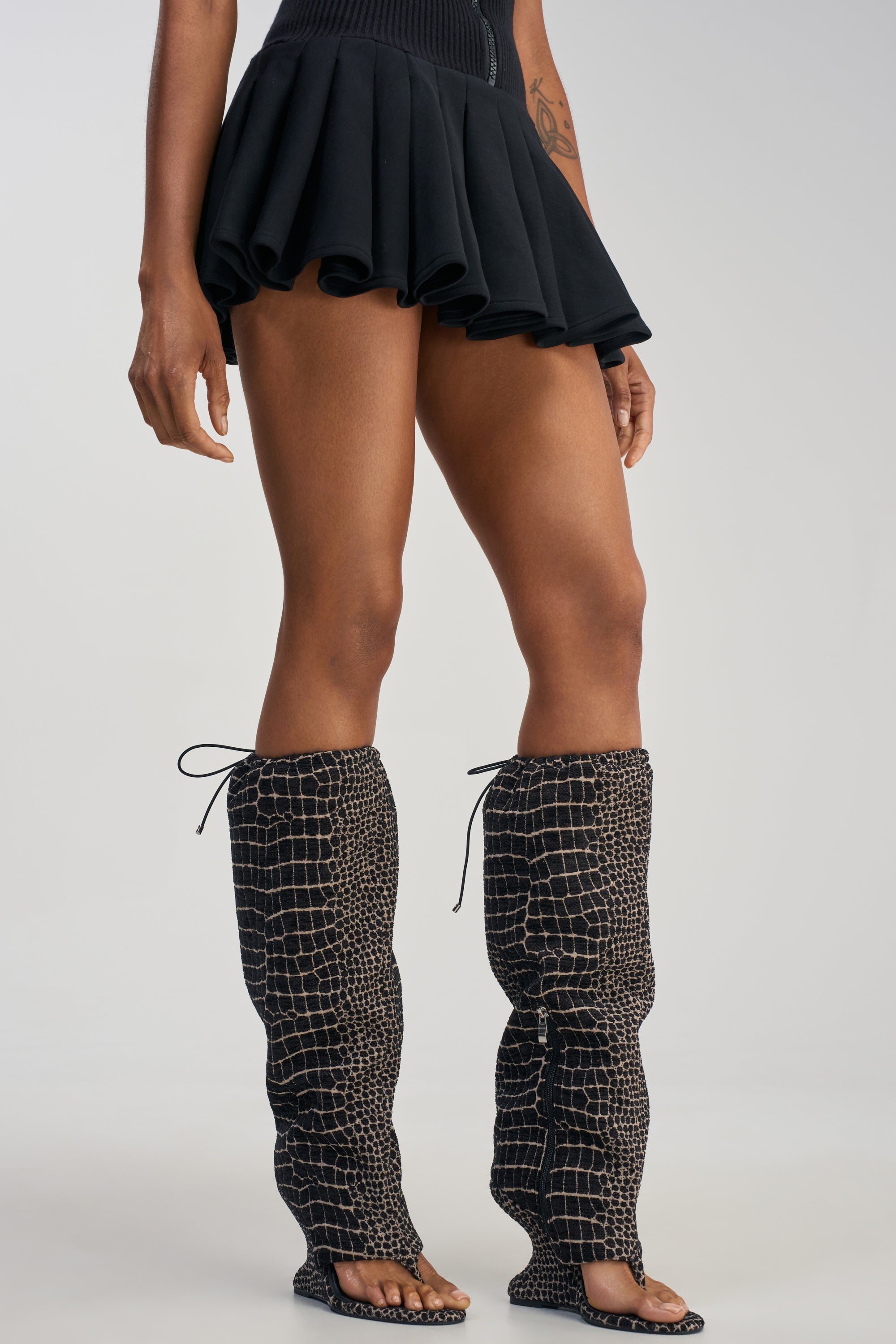A person wearing a short black skirt and the AZALEA WANG POET BLACK AND WHITE SNAKE PATTERN WEDGE SANDAL, featuring open toes and a knee-high shaft with a black-and-white textured snake pattern that covers their legs up to the knees. The background is plain and light-colored.