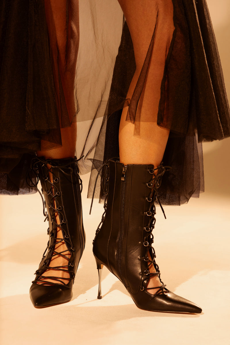 Someone wears the AZALEA WANG PONCIA BLACK LACE UP BLACK BOOTIE with pointed toes and stiletto heels, paired with a sheer black tulle skirt. Dramatic lighting adds to the stylish overall look.
