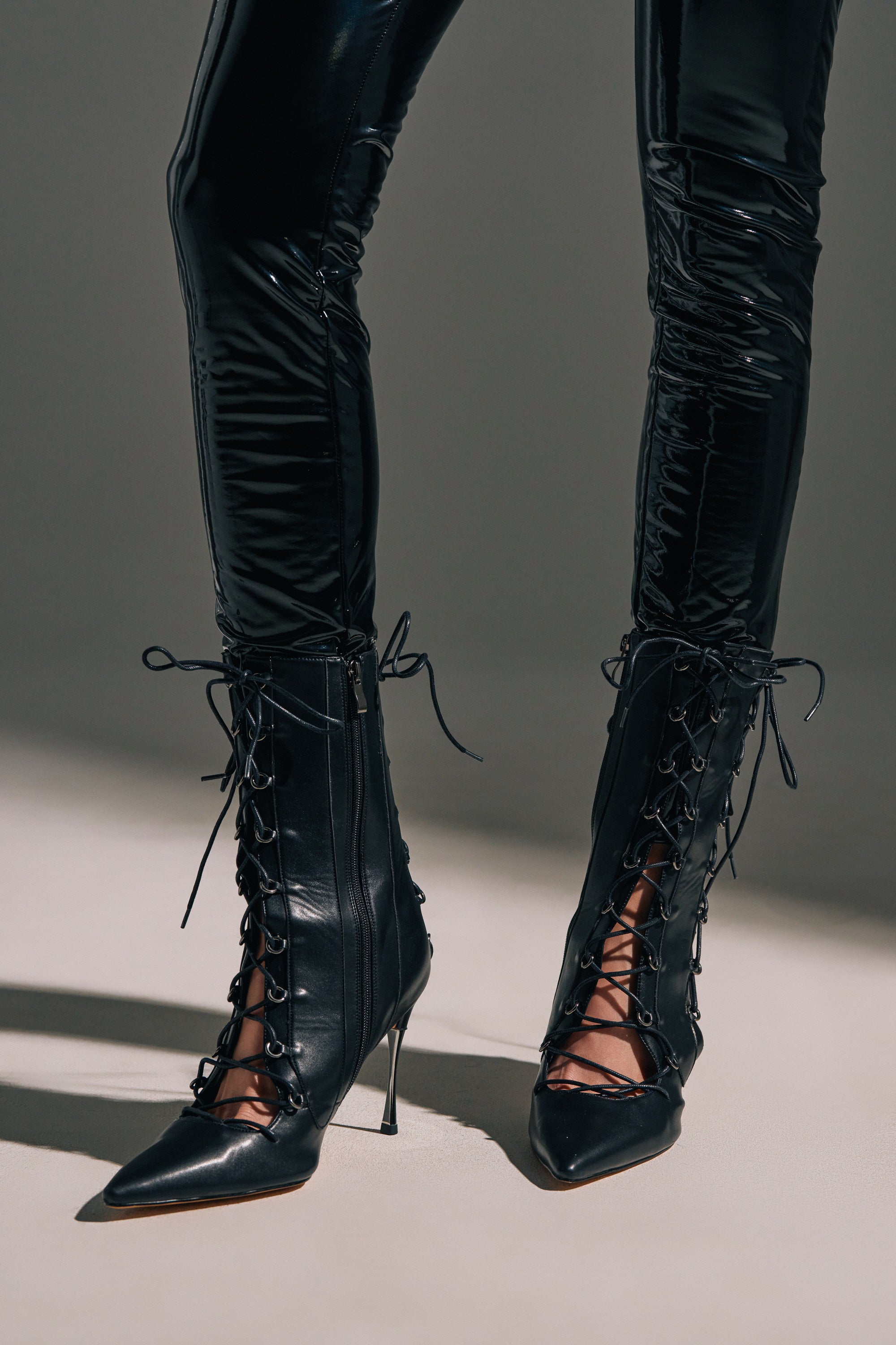 Wearing shiny black vinyl pants, a person stands on a light surface, showcasing the chic allure of each pointed-toe stiletto heel on the AZALEA WANG PONCIA BLACK LACE UP BLACK BOOTIE.