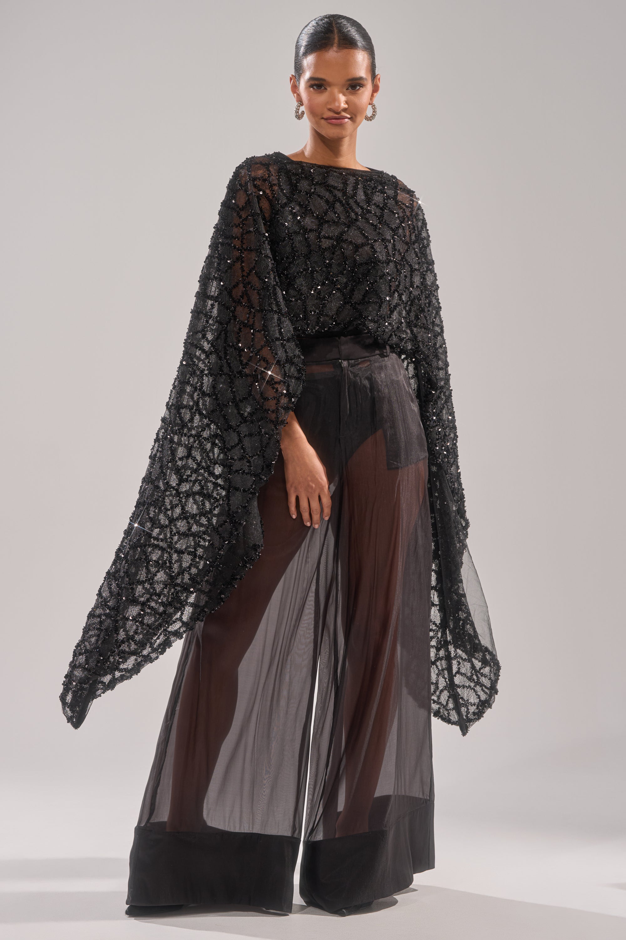 A woman models the SHINE AWAY MESH BLOUSE, featuring sheer mesh fabric, batwing sleeves, and sequin details, paired with wide-leg pants. She stands confidently against a plain light background.