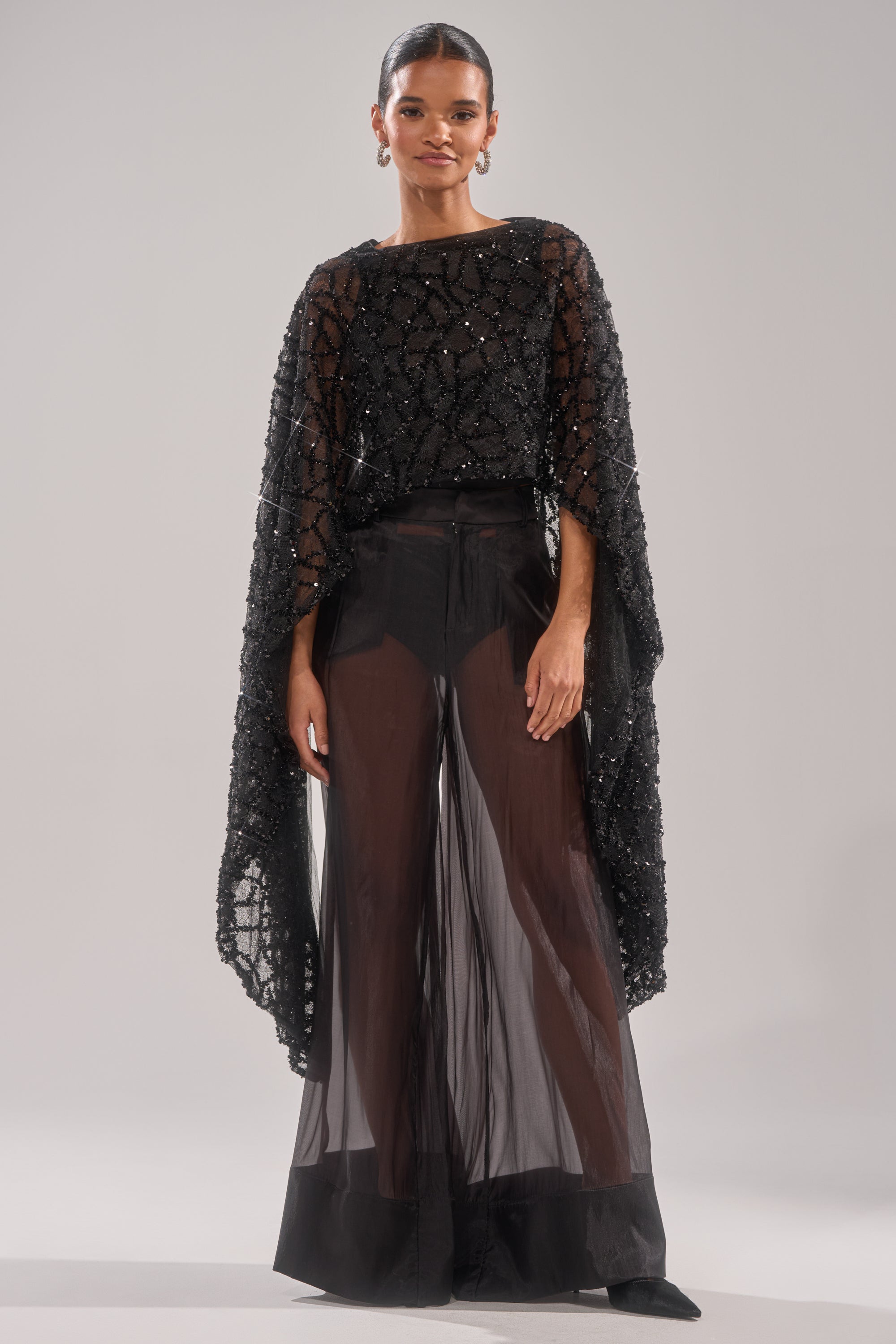 A woman wears the SHINE AWAY MESH BLOUSE with a sheer black sequin cape and wide-leg pants, hair pulled back and hoop earrings, standing against a light background.