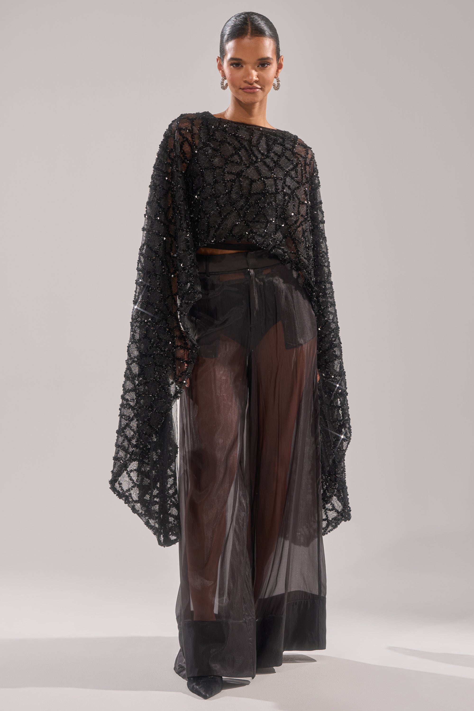 Wearing the SHINE AWAY MESH BLOUSE with intricate beading and dramatic batwing sleeves, she pairs it with high-waisted sheer black pants and pointed shoes, posing against a simple, light background.