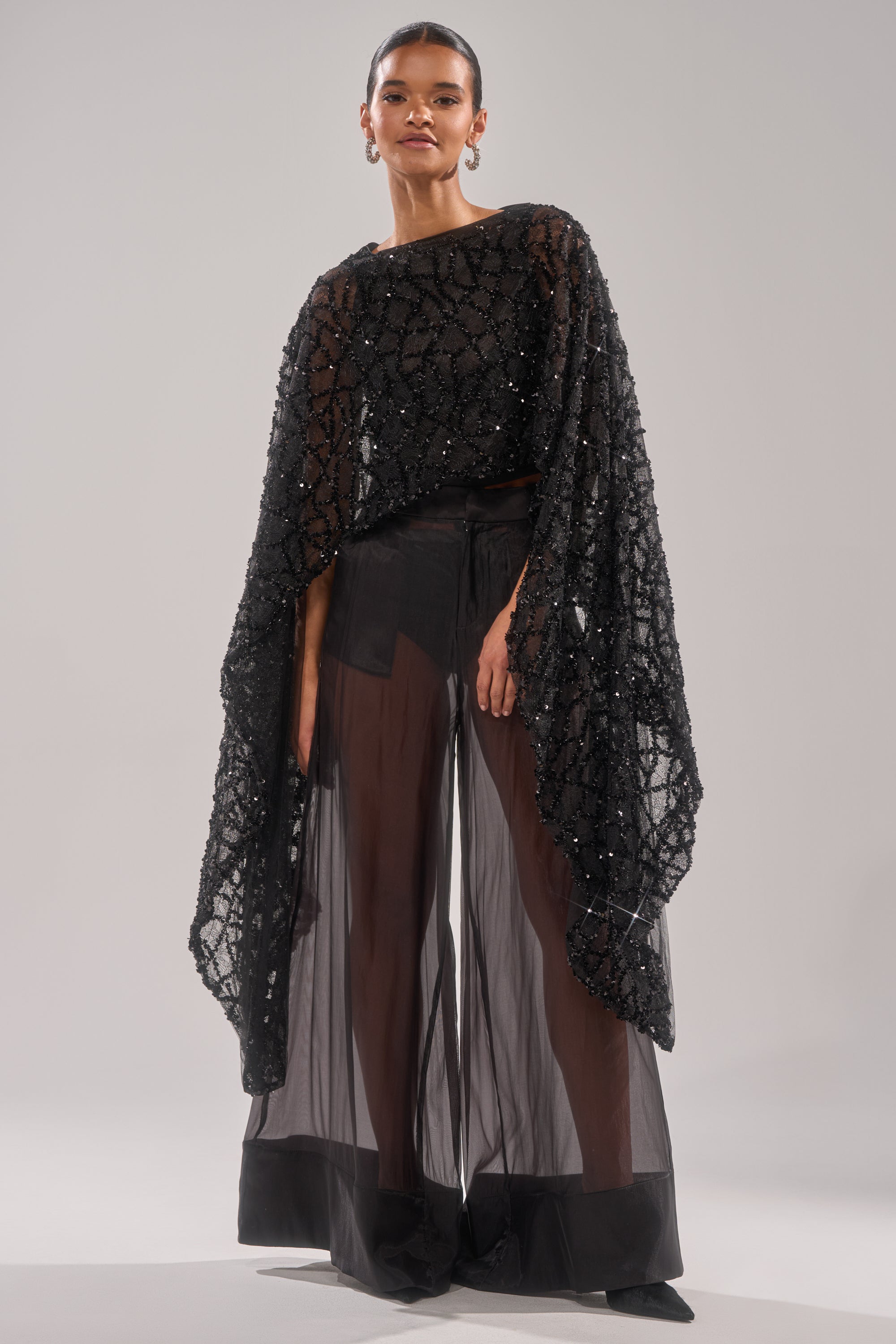 A woman stands confidently in the SHINE AWAY MESH BLOUSE, featuring sheer black mesh with batwing sleeves and intricate beading, paired with wide-legged transparent pants and a black top against a plain light background.