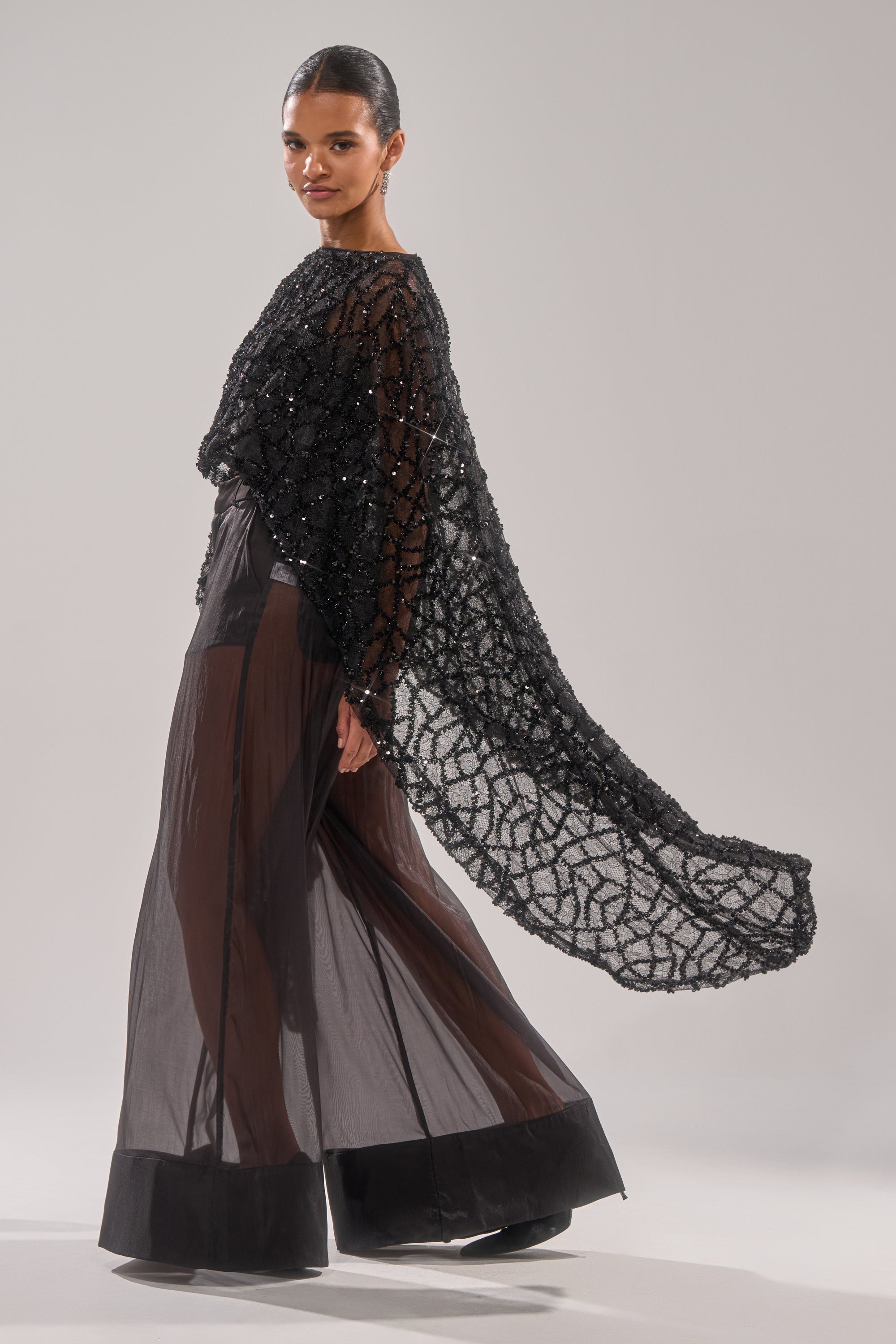 A person wears the SHINE AWAY MESH BLOUSE, a sheer black top with batwing sleeves, paired with wide-leg pants and a beaded geometric cape draped from the shoulders. The background is plain and light-colored.