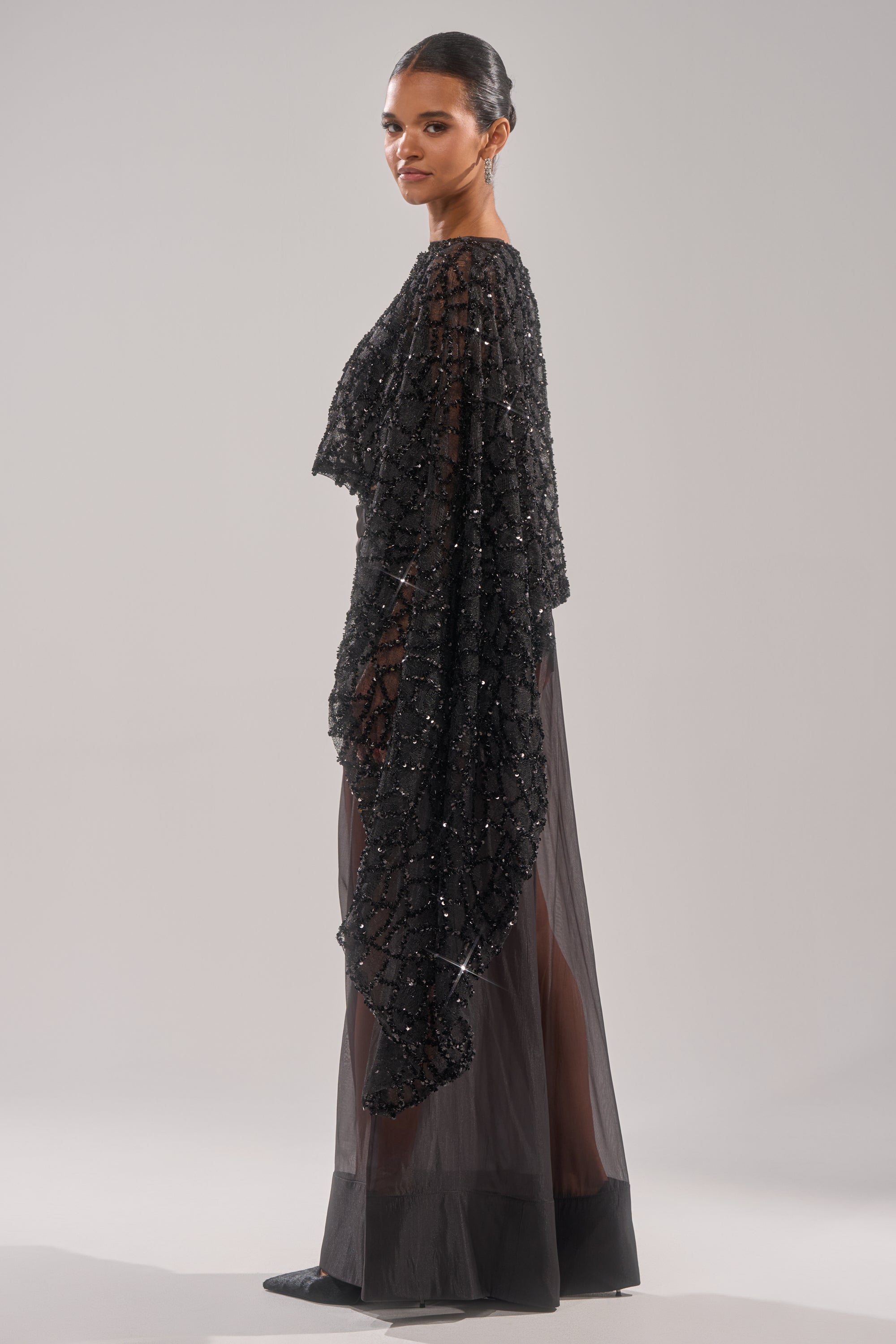 A woman stands sideways against a light gray background, wearing the SHINE AWAY MESH BLOUSE—a sheer black design paired with a long beaded cape. Her hair is tied back, and the floor-length dress showcases her legs beneath the mesh fabric.
