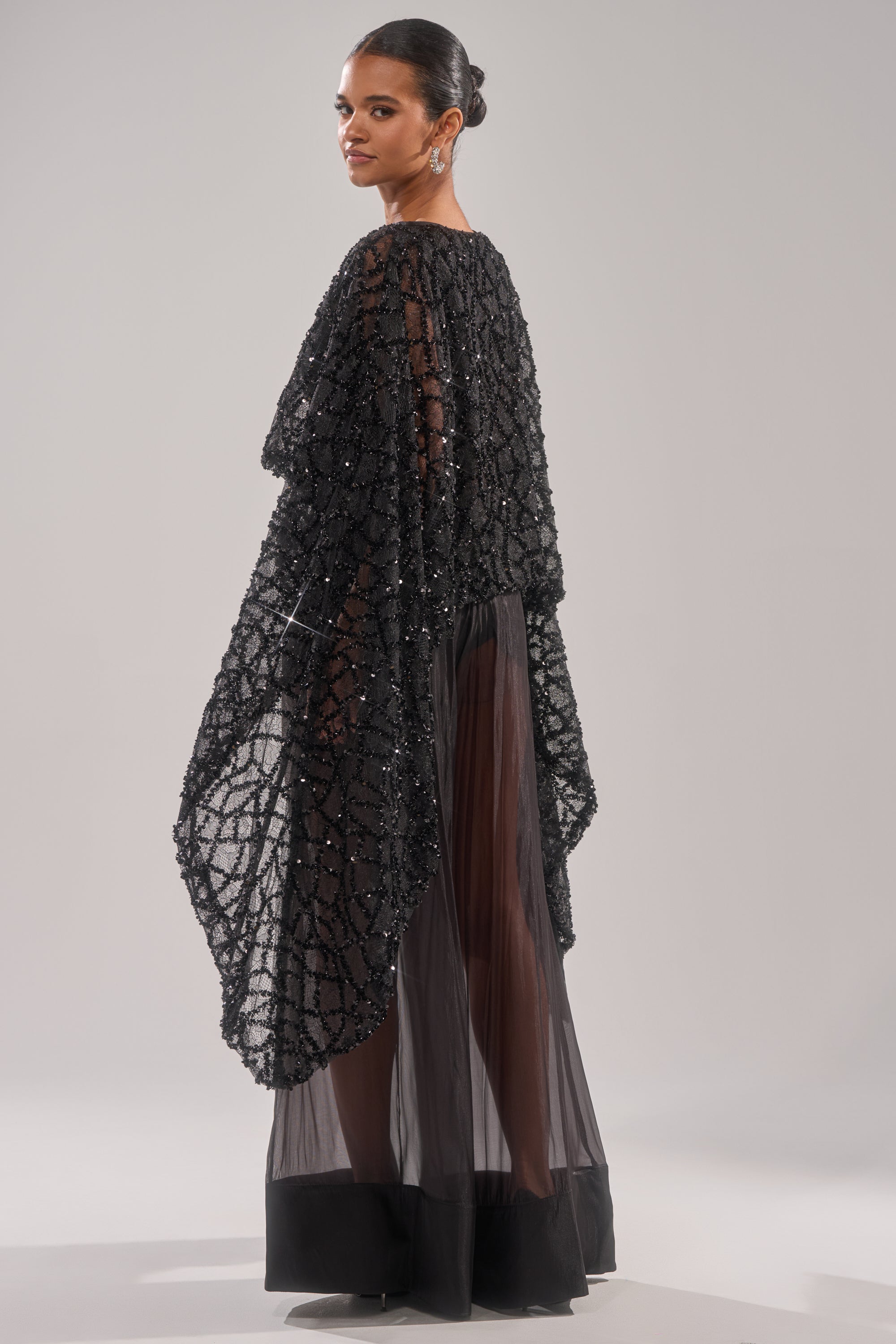 A woman stands sideways in the SHINE AWAY MESH BLOUSE, featuring sheer black fabric with bold batwing sleeves and textured beaded detailing. Her hair is styled in a sleek bun against a plain, light background.