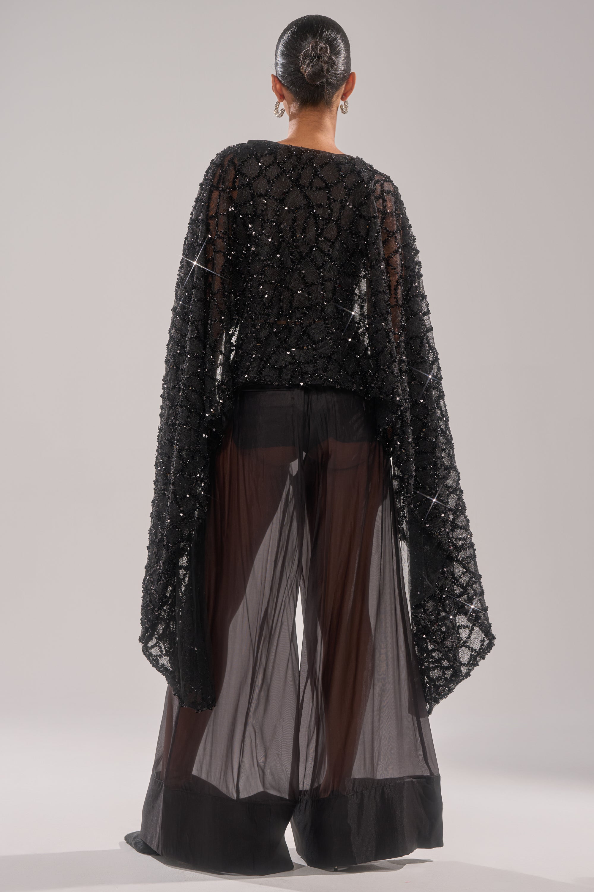 A person with hair in a sleek bun is shown from behind, wearing the SHINE AWAY MESH BLOUSE and wide, sheer black pants over black shorts, standing against a plain light background.