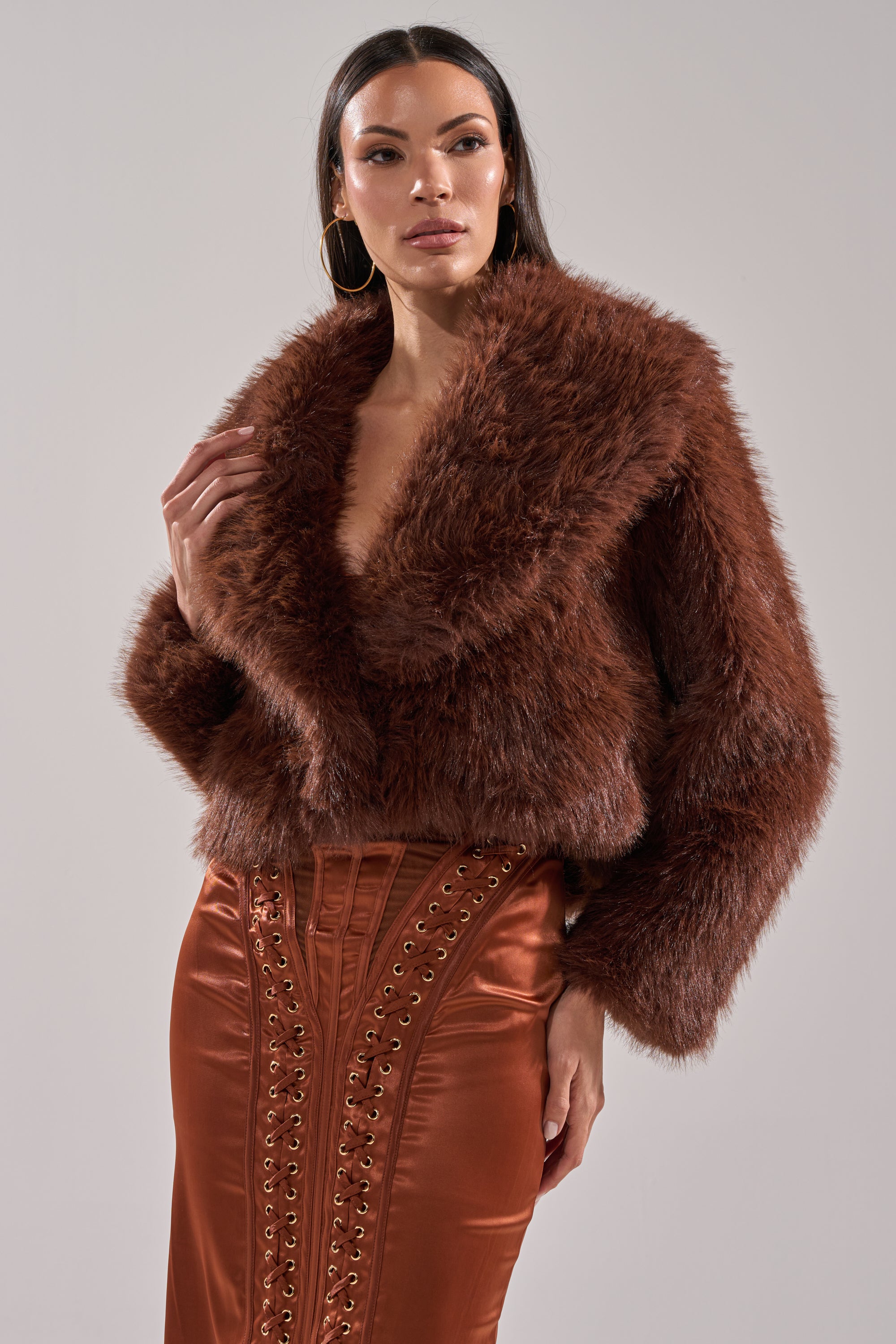 A woman in the POPPY CROPPED FAUX FUR IN BROWN jacket and a rust satin lace-up skirt stands against a plain background, looking confidently to the side.