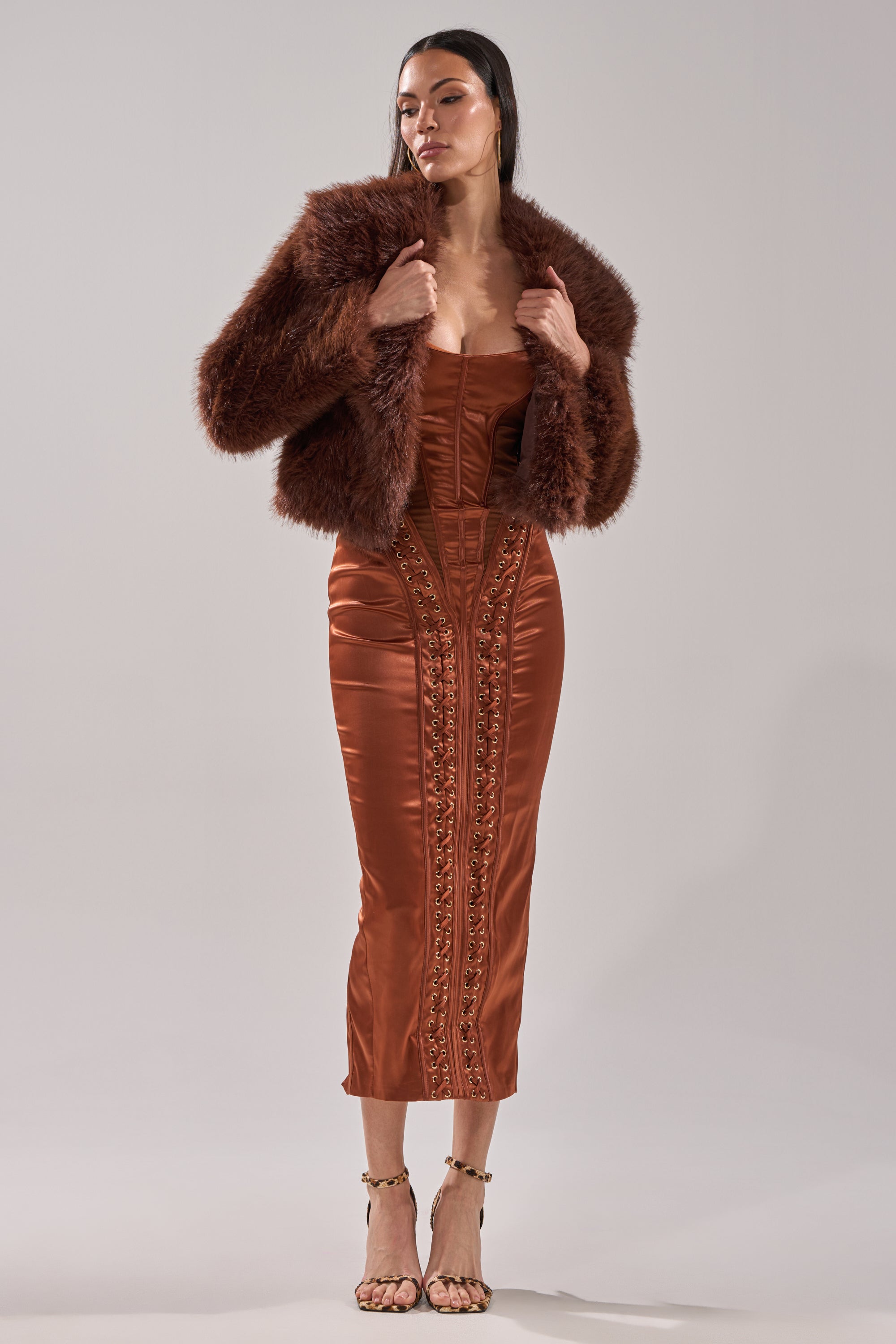 A woman wears a rust satin midi dress with ornate details, layered with the POPPY CROPPED FAUX FUR IN BROWN. She poses holding the jacket's collar and completes her look with strappy high-heeled sandals.