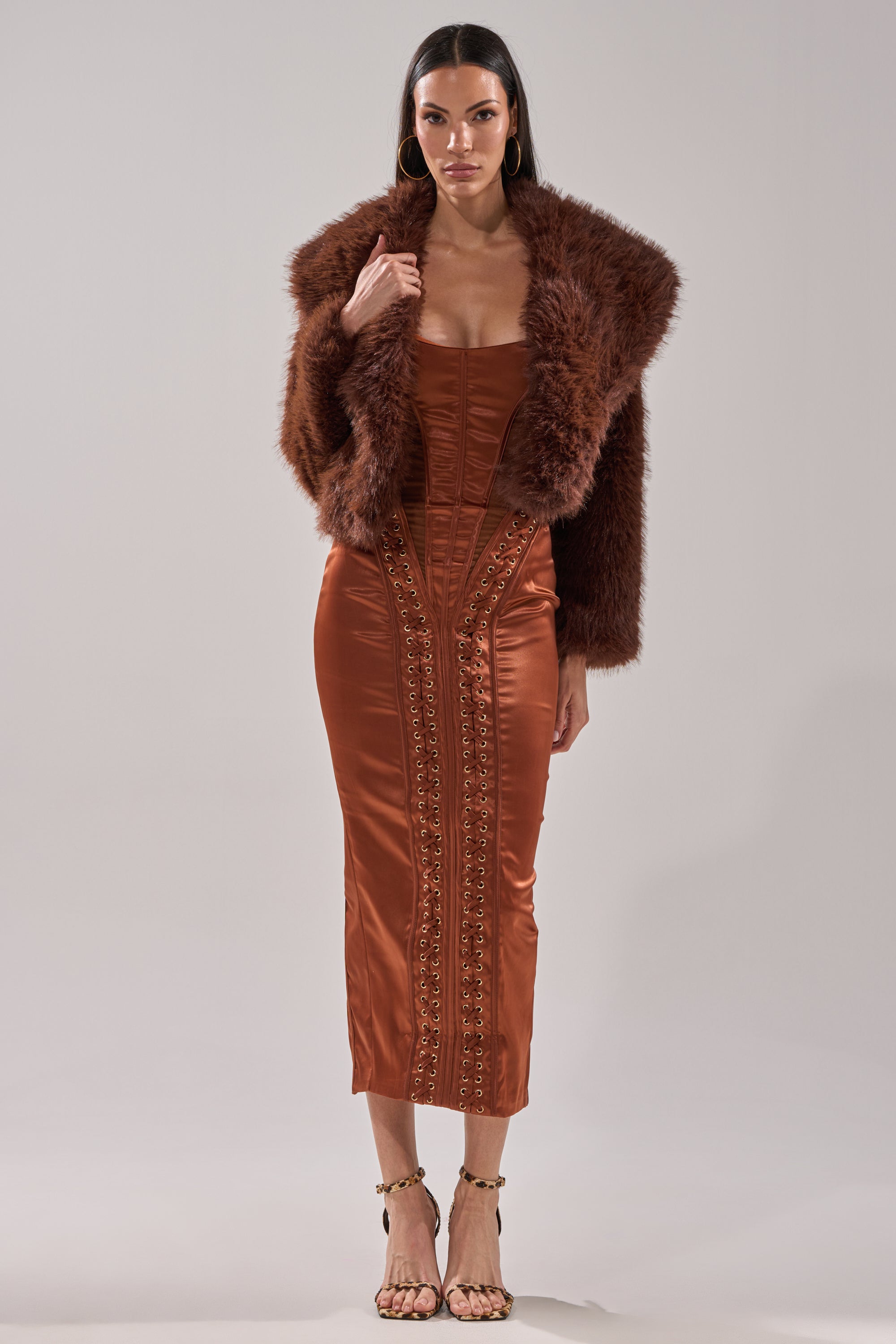 A woman wears the POPPY Cropped Faux Fur in Brown over a rust satin midi dress with decorative stitching and strappy heels, standing against a plain light gray background.