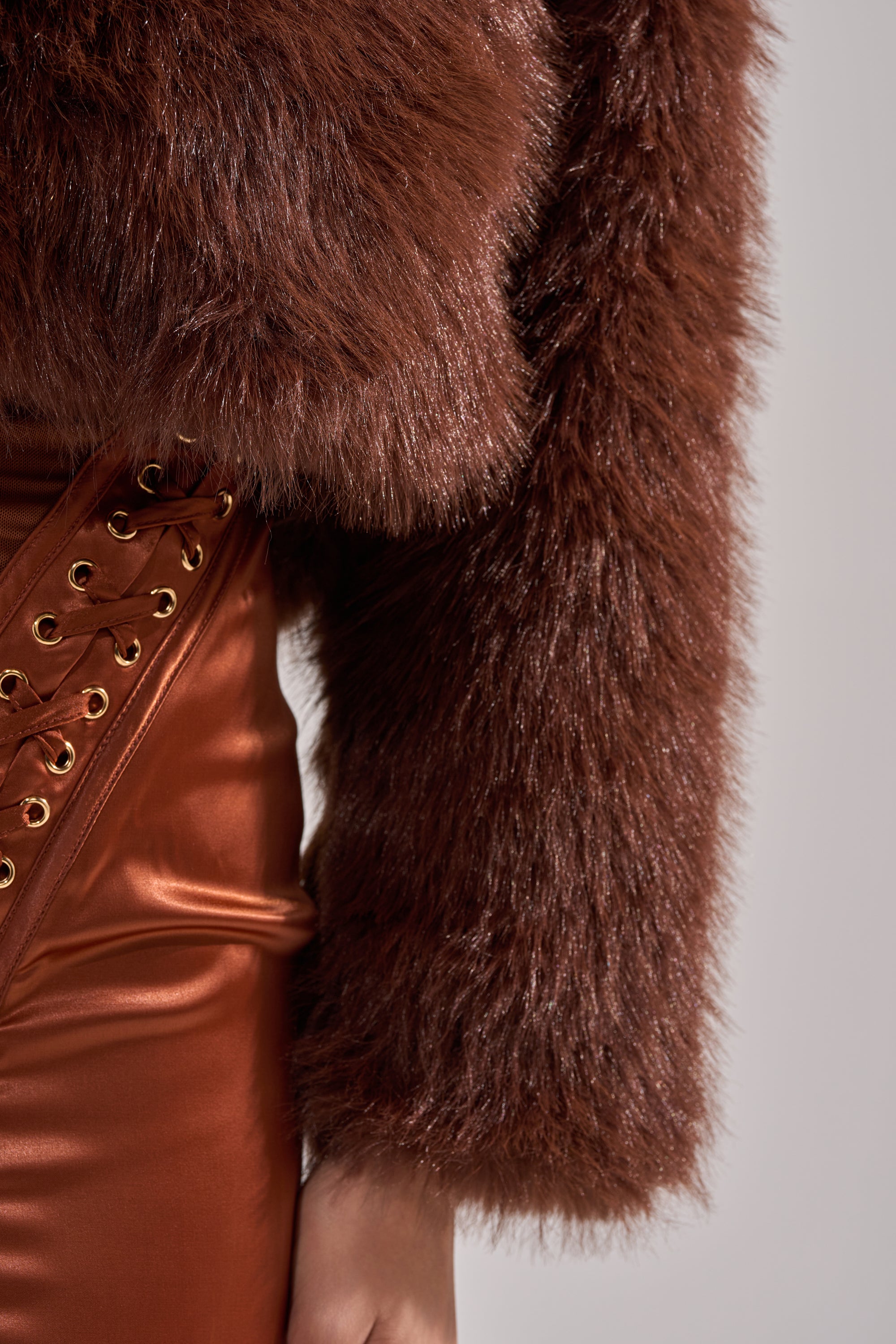Wearing the POPPY Cropped Faux Fur in Brown and a shiny brown leather skirt with gold eyelet lace-up detail. Only the torso and part of one arm are visible.