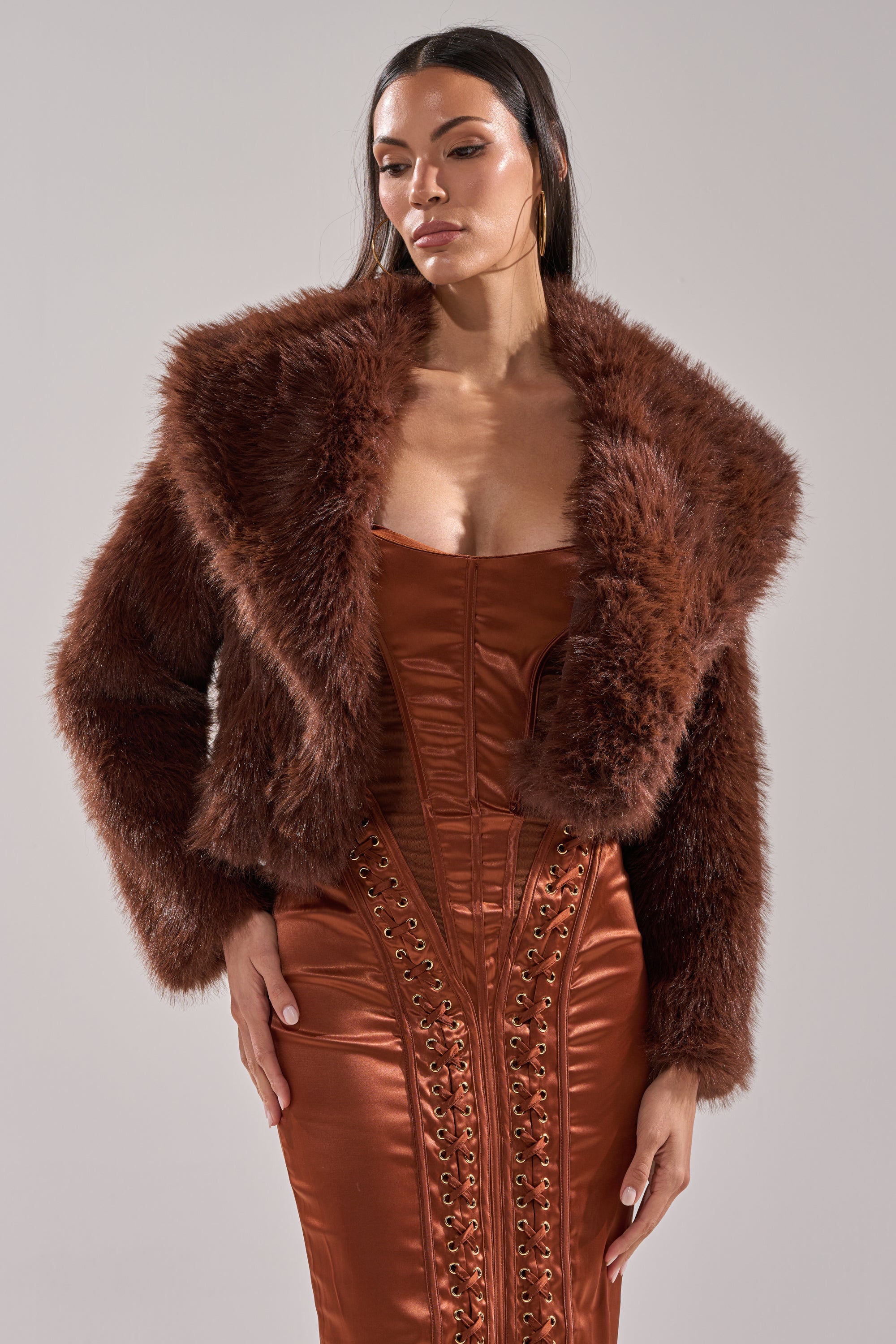 A woman wears the POPPY CROPPED FAUX FUR IN BROWN with an oversized shawl collar over a shiny rust dress with lace-up details, standing against a plain light gray background.