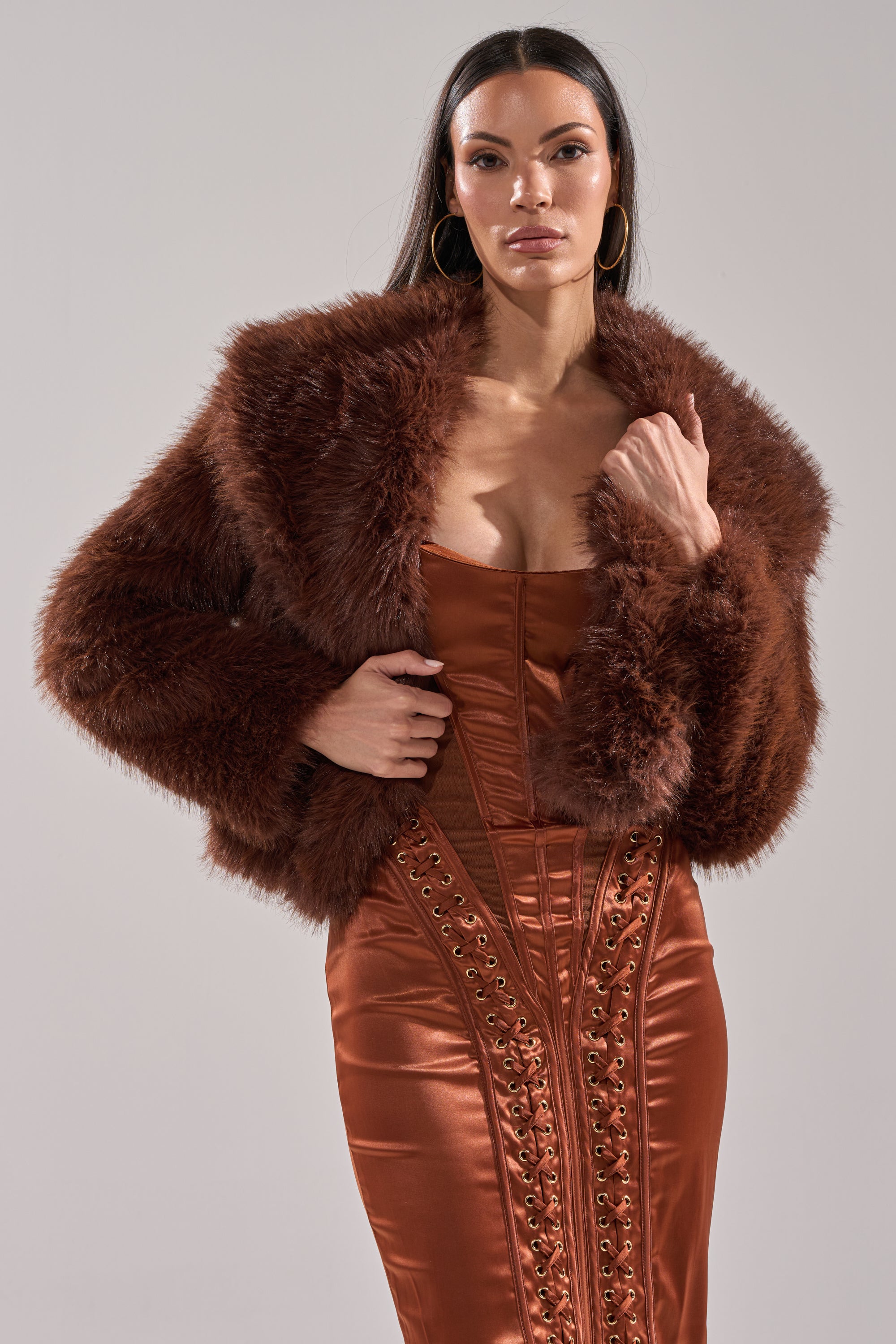 A woman poses confidently against a gray backdrop, wearing the POPPY Cropped Faux Fur in Brown over a shiny, rust-colored corset dress with decorative lacing.