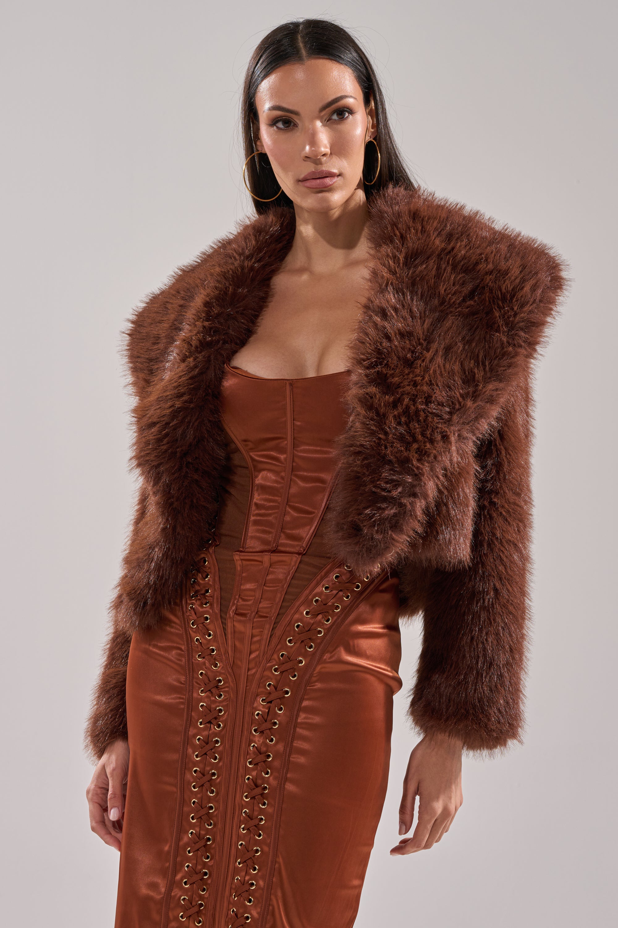 A woman wears the POPPY CROPPED FAUX FUR IN BROWN over a copper satin lace-up dress with large gold hoops, posing against a plain light background.