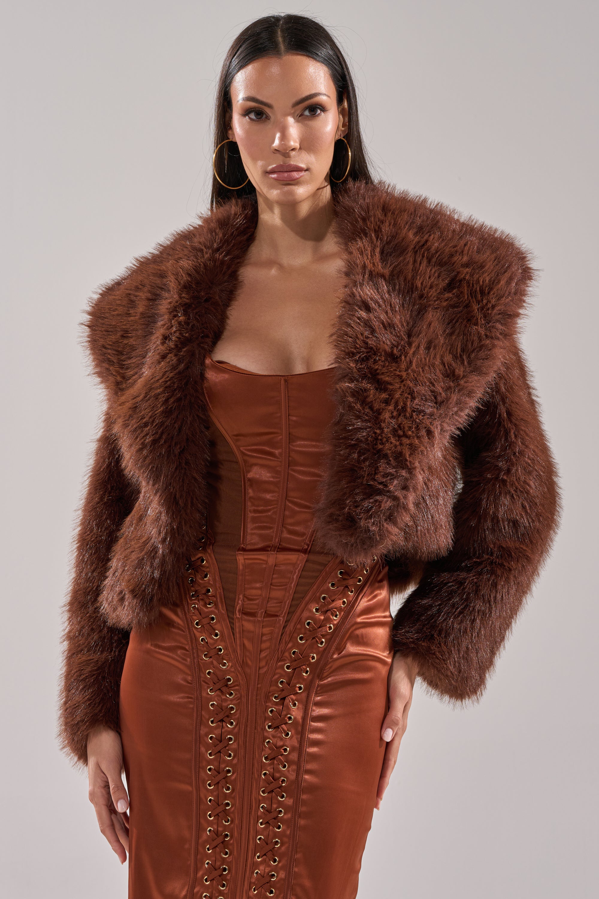 A woman models the POPPY CROPPED FAUX FUR IN BROWN over a fitted, rust satin dress with corset-style lacing, standing against a simple gray backdrop.