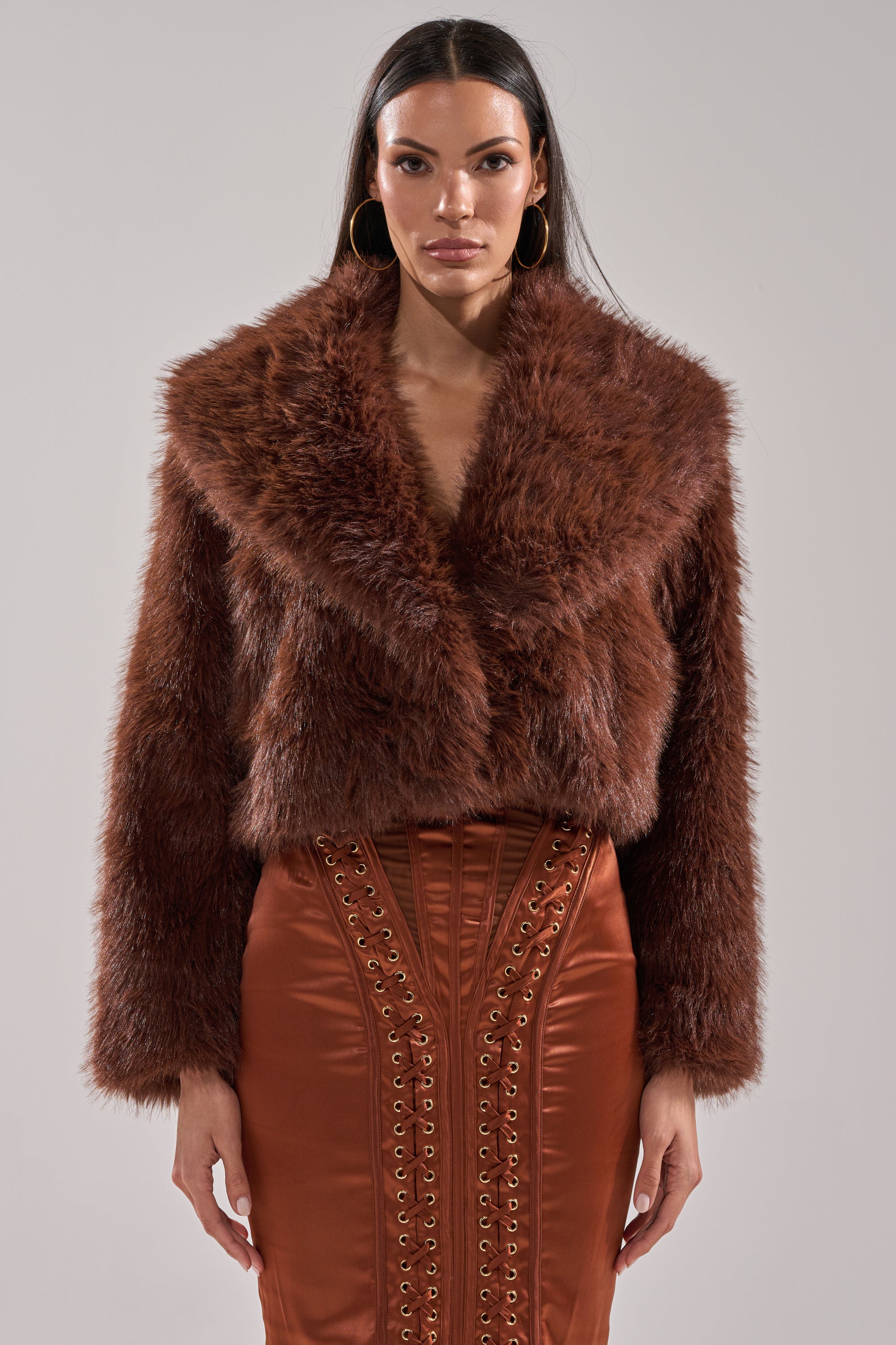 A woman with long dark hair and large hoop earrings faces forward, wearing the POPPY CROPPED FAUX FUR IN BROWN over a shiny rust high-waisted skirt with lace-up details.