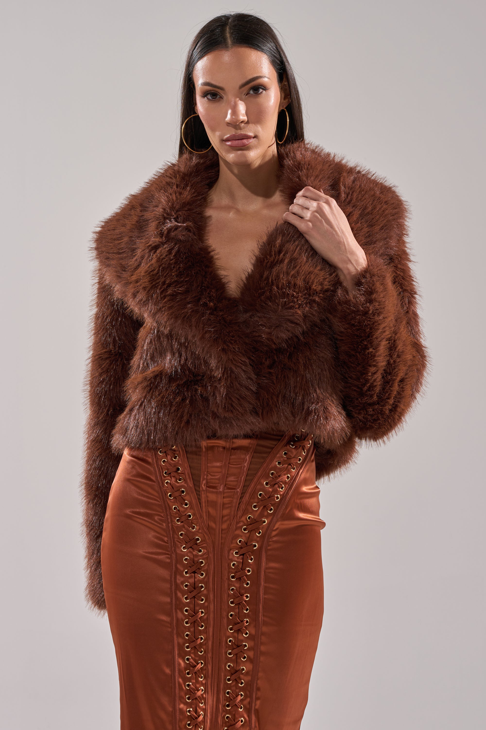 A woman wears the POPPY CROPPED FAUX FUR IN BROWN jacket with a high-waisted burnt orange satin skirt with lace-up details, standing against a plain light gray background.