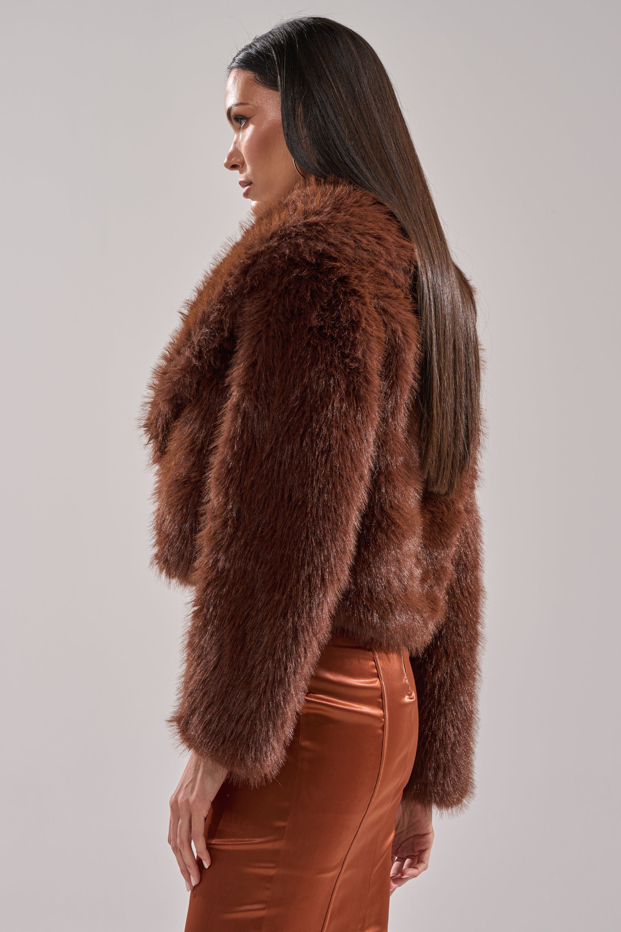 A woman with long, straight brown hair wears the POPPY CROPPED FAUX FUR IN BROWN, paired with a shiny, high-waisted copper skirt, standing sideways and looking forward against a plain light background.