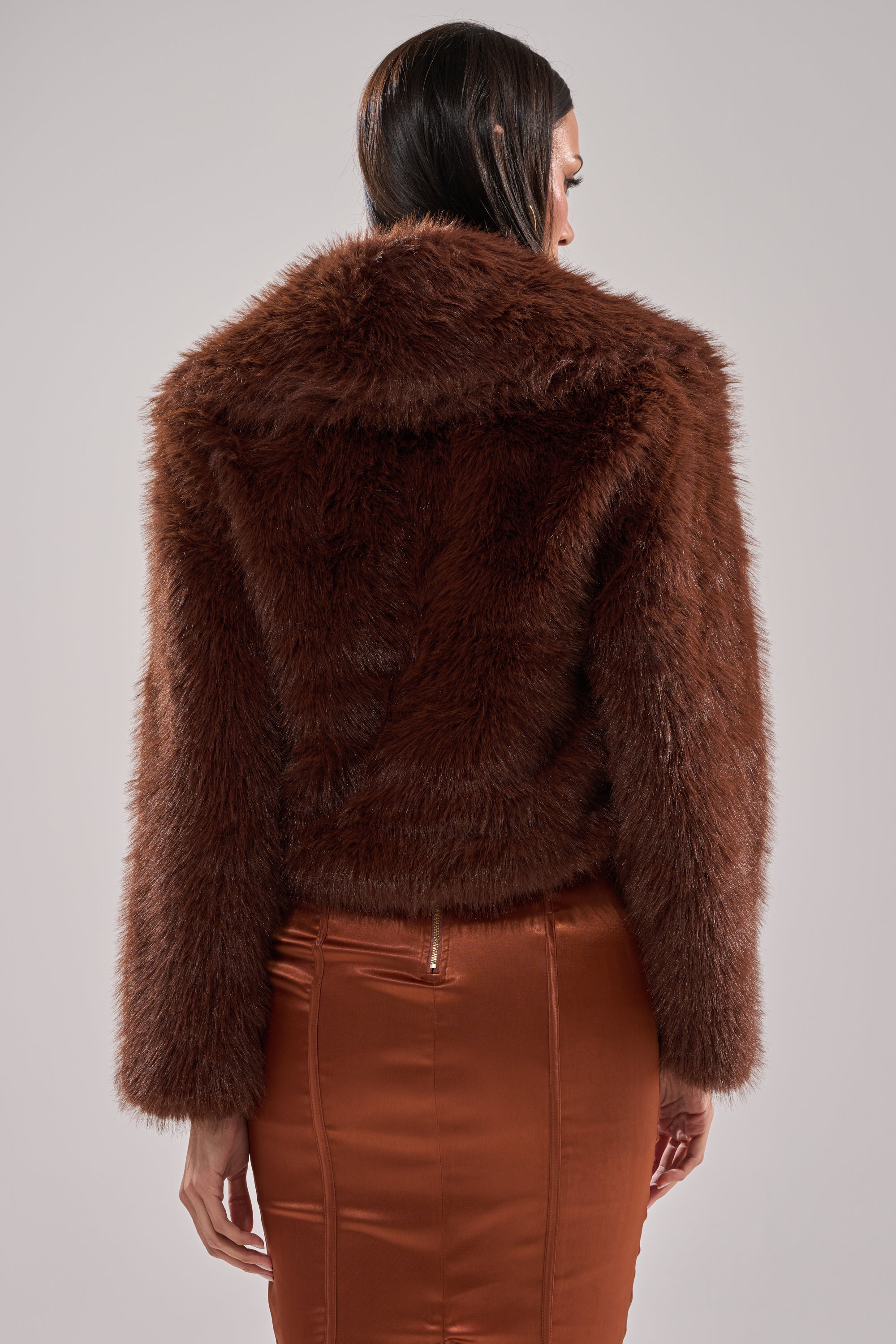 A person faces away from the camera, wearing the POPPY CROPPED FAUX FUR IN BROWN coat and a shiny copper skirt, set against a plain light gray background.