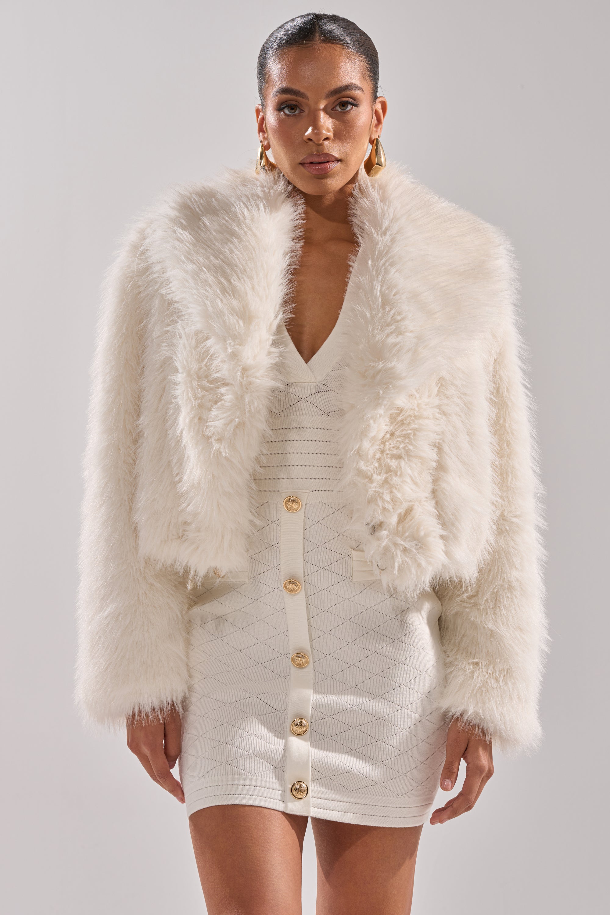 A woman wears the POPPY Cropped Faux Fur in White over a fitted white dress with a deep V-neck and gold buttons, accessorized with gold hoop earrings, against a plain light background.