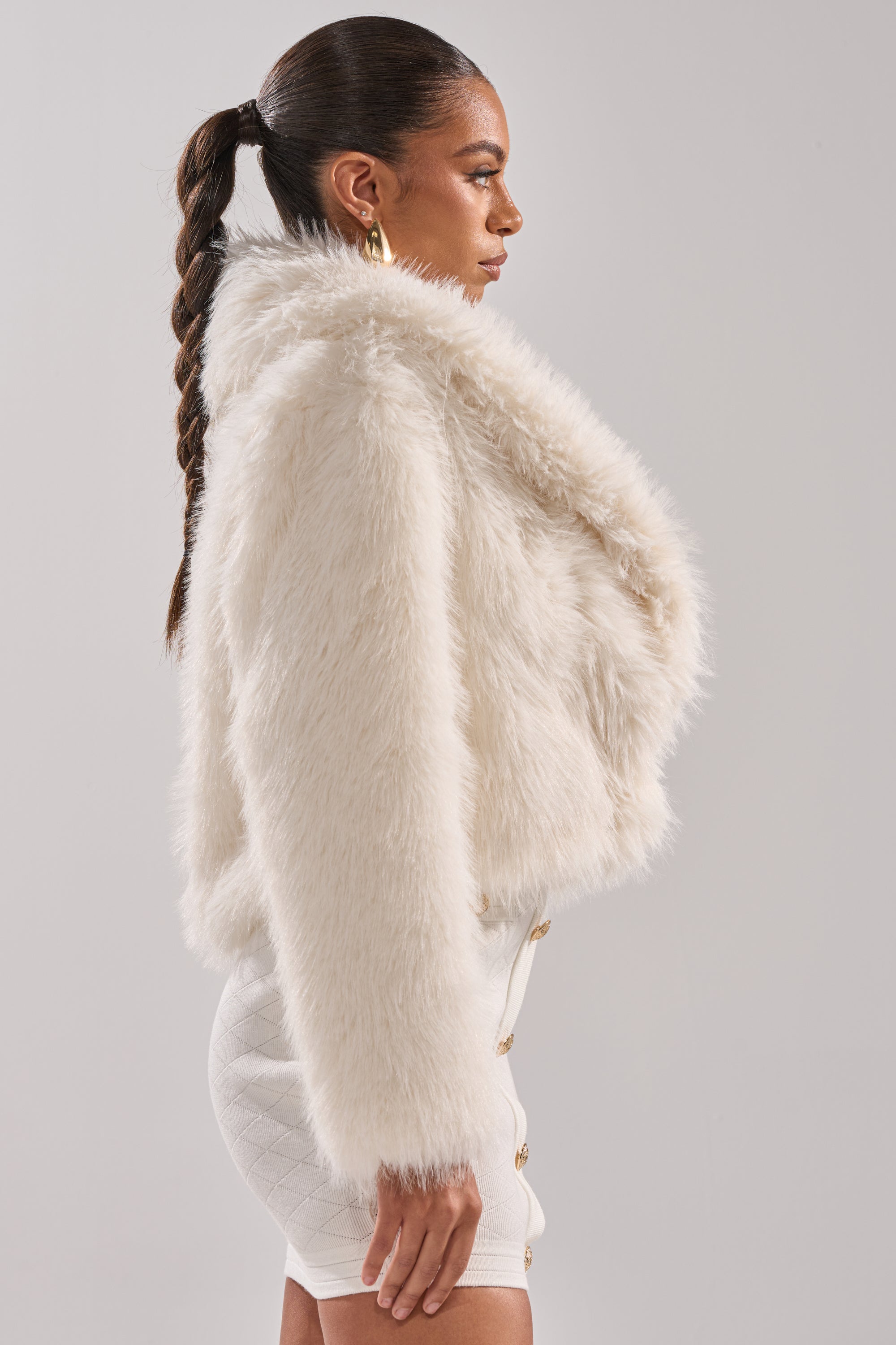 Woman in profile wears the AKIRA Label POPPY CROPPED FAUX FUR IN WHITE jacket over a white fitted skirt with gold buttons and gold hoop earrings. Her long braided hair stands out against a plain light gray background.