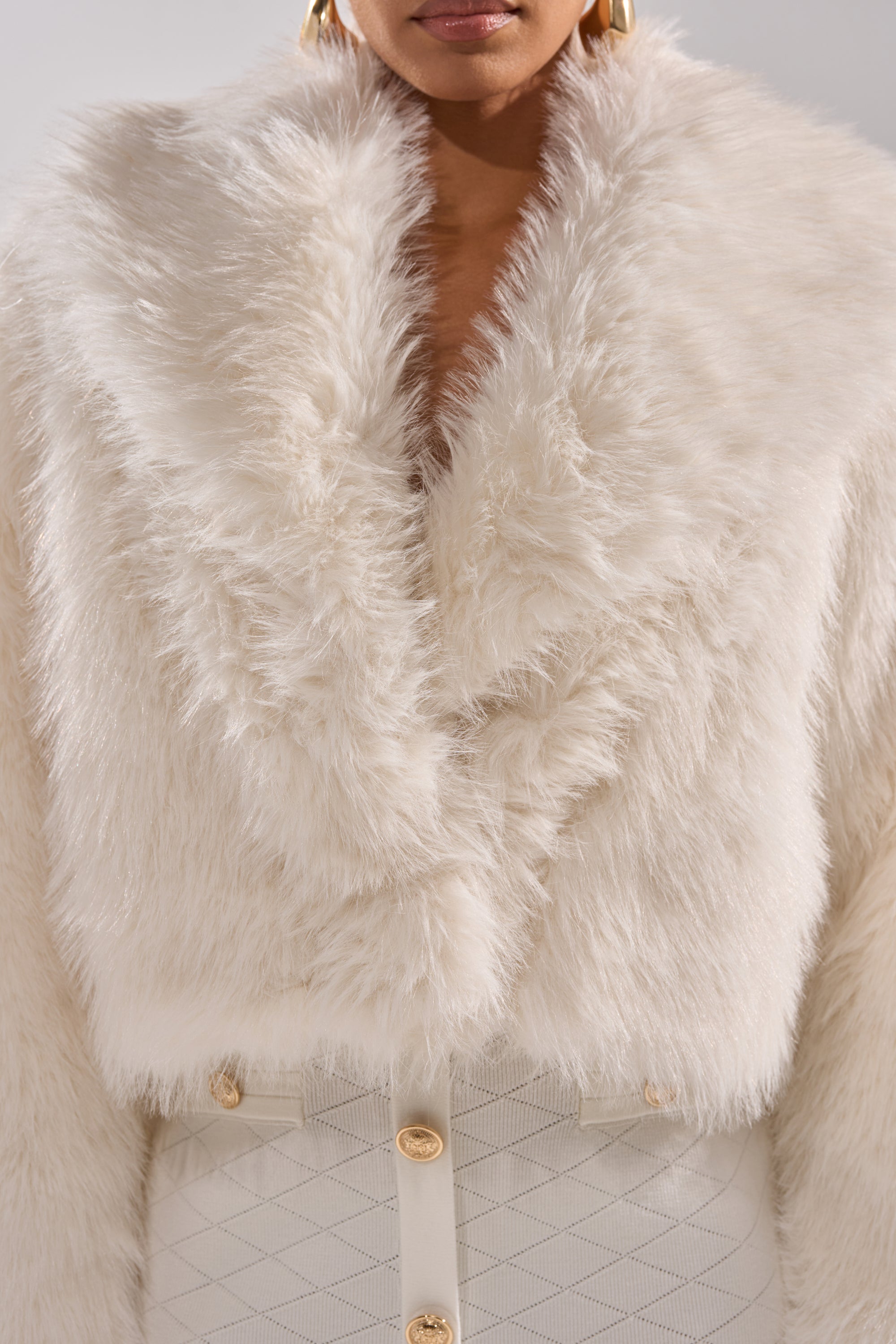 A woman wears the POPPY CROPPED FAUX FUR IN WHITE, a luxurious faux fur coat with a wide collar, paired with a high-waisted textured white skirt with gold buttons. Her gold hoop earrings and the lower half of her face are visible.