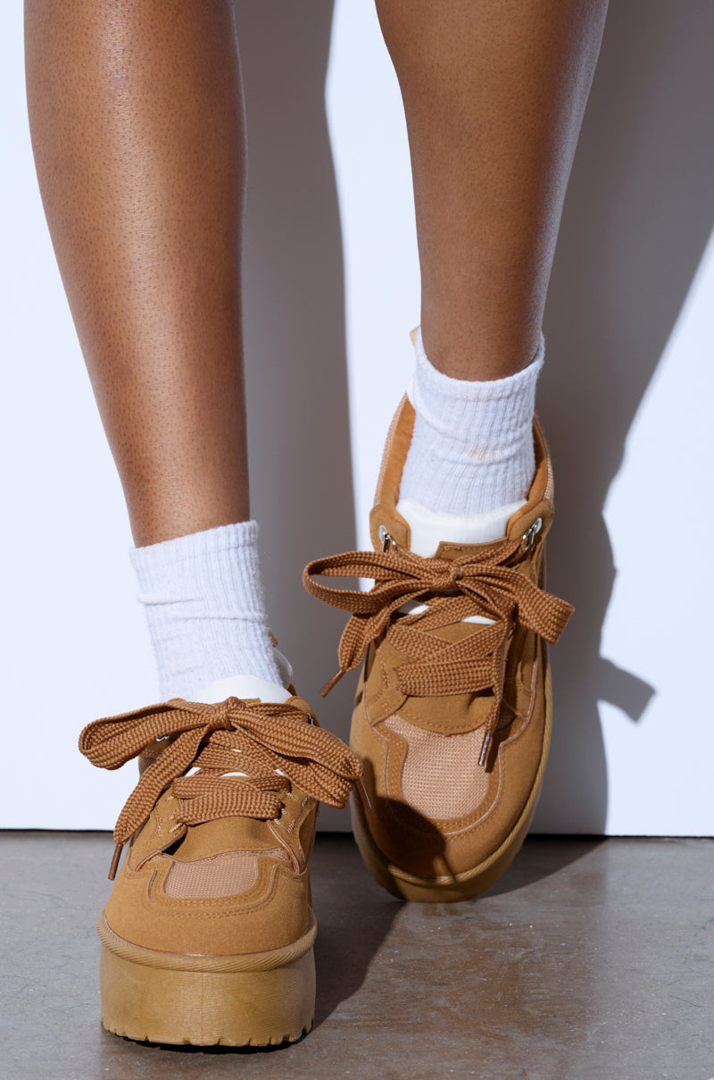 A person wears the POPULARITY CHESTNUT FLATFORM SNEAKER, featuring matching tan laces, chestnut suede upper, and thick soles, with white socks on a concrete surface. Their legs cast a soft shadow on the white background.