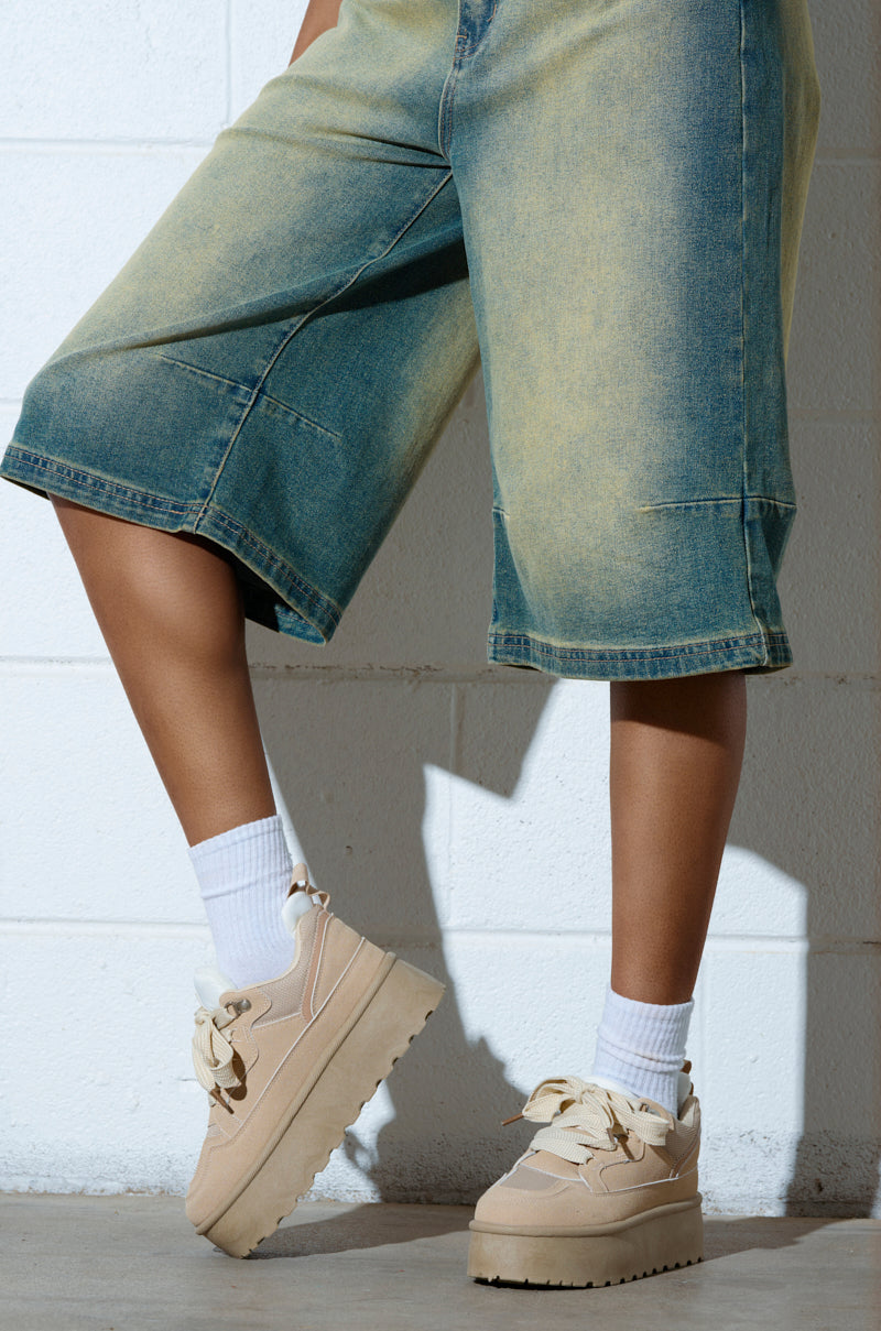 Against a white brick wall, a person in faded denim shorts and white socks highlights their chic POPULARITY SAND FLATFORM SNEAKER with an almond toe. One leg is slightly bent, emphasizing the stylish footwear and casual slip-on fit. 1