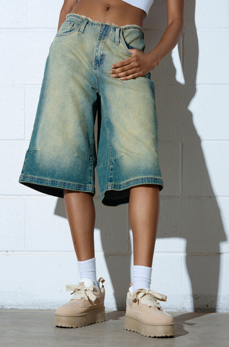 A person in wide, knee-length denim shorts, a white crop top, and white socks with beige POPULARITY SAND FLATFORM SNEAKER stands against a white brick wall with one hand in their pocket. 2