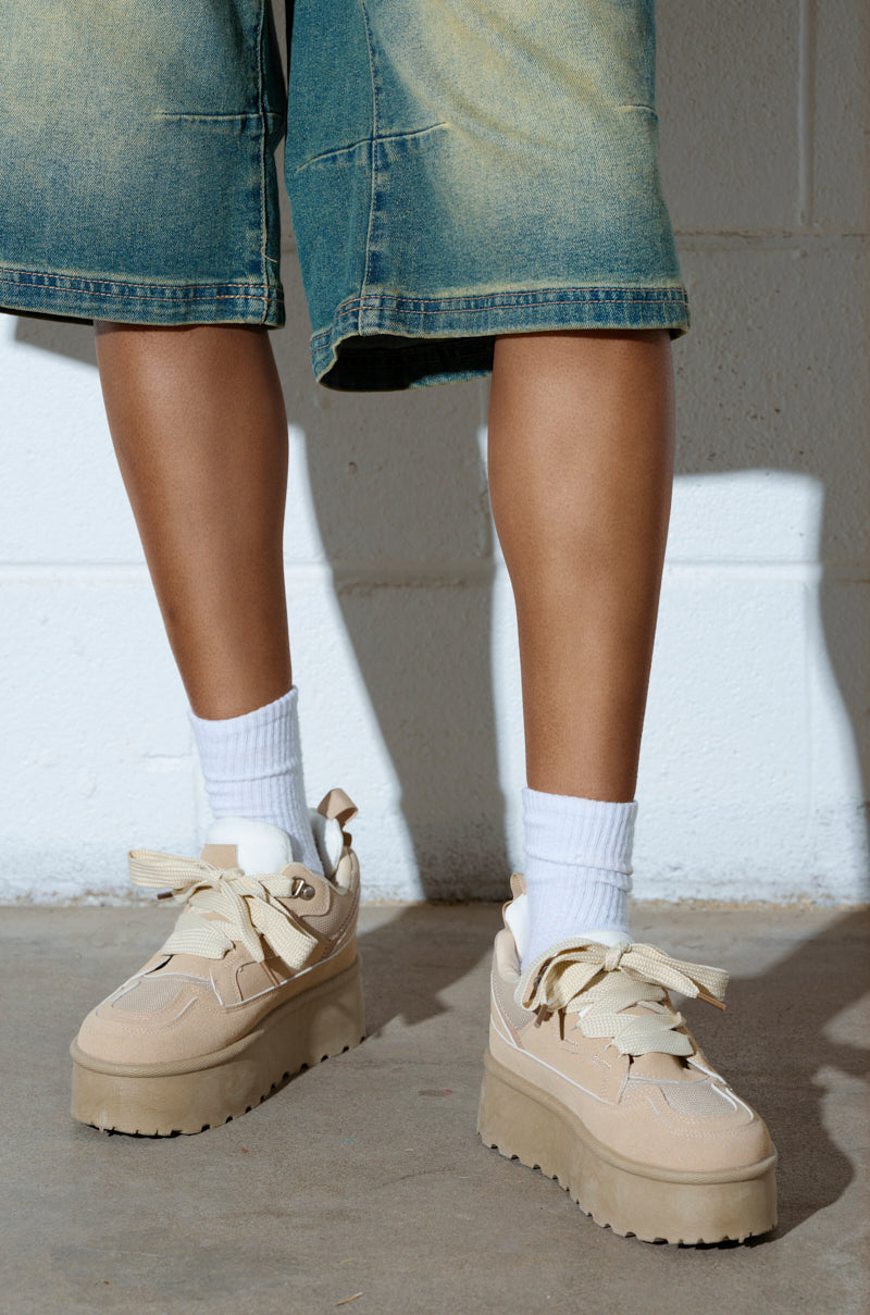 Sporting POPULARITY SAND FLATFORM SNEAKER in light beige with white socks, a person stands on a concrete floor dressed in faded denim shorts against a white brick wall. 0