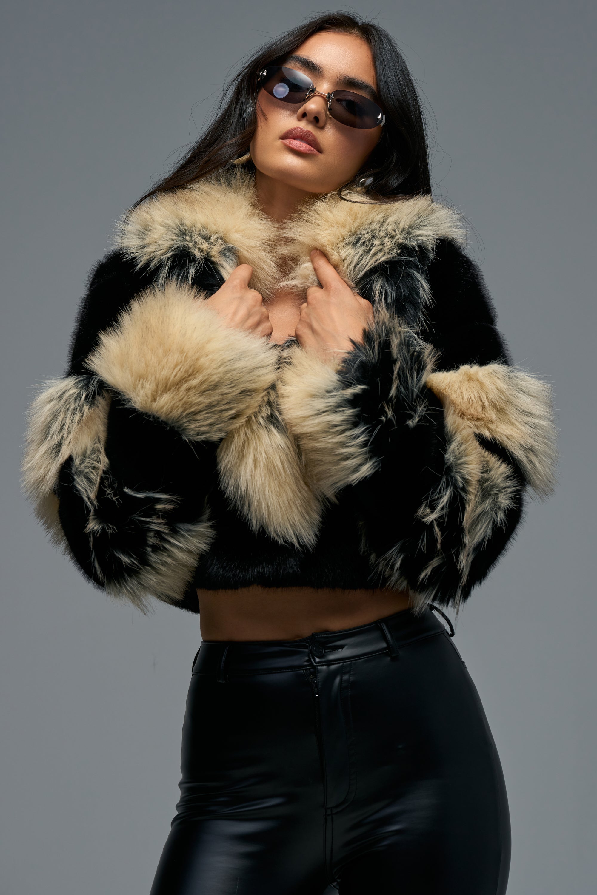 A woman wearing sunglasses, the PORTIA CROPPED FAUX FUR IN BLACK BROWN jacket, and black leather pants poses confidently against a plain gray background.