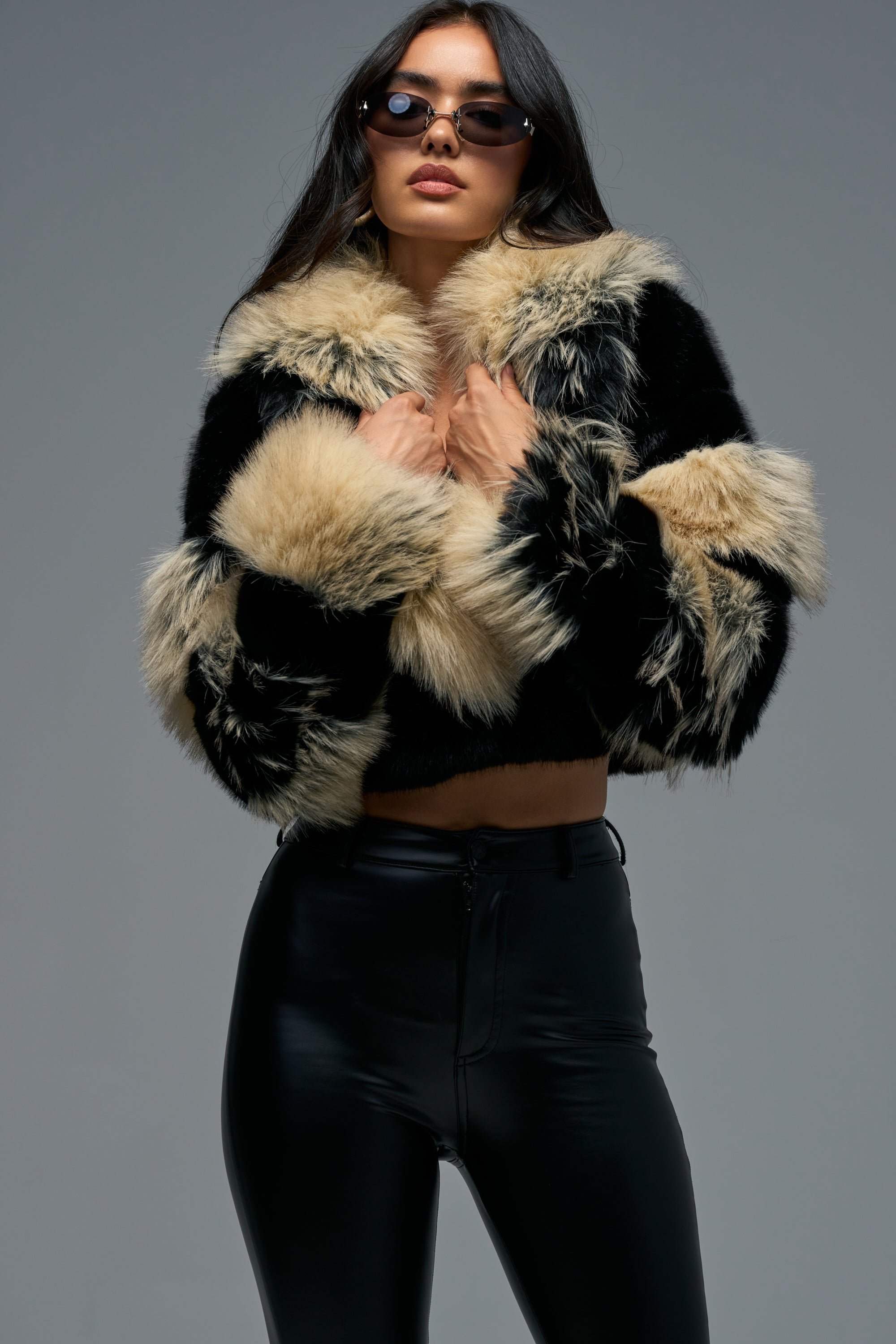 A woman stands confidently against a plain gray background, wearing the PORTIA CROPPED FAUX FUR IN BLACK BROWN jacket with black high-waisted leather pants and sunglasses.