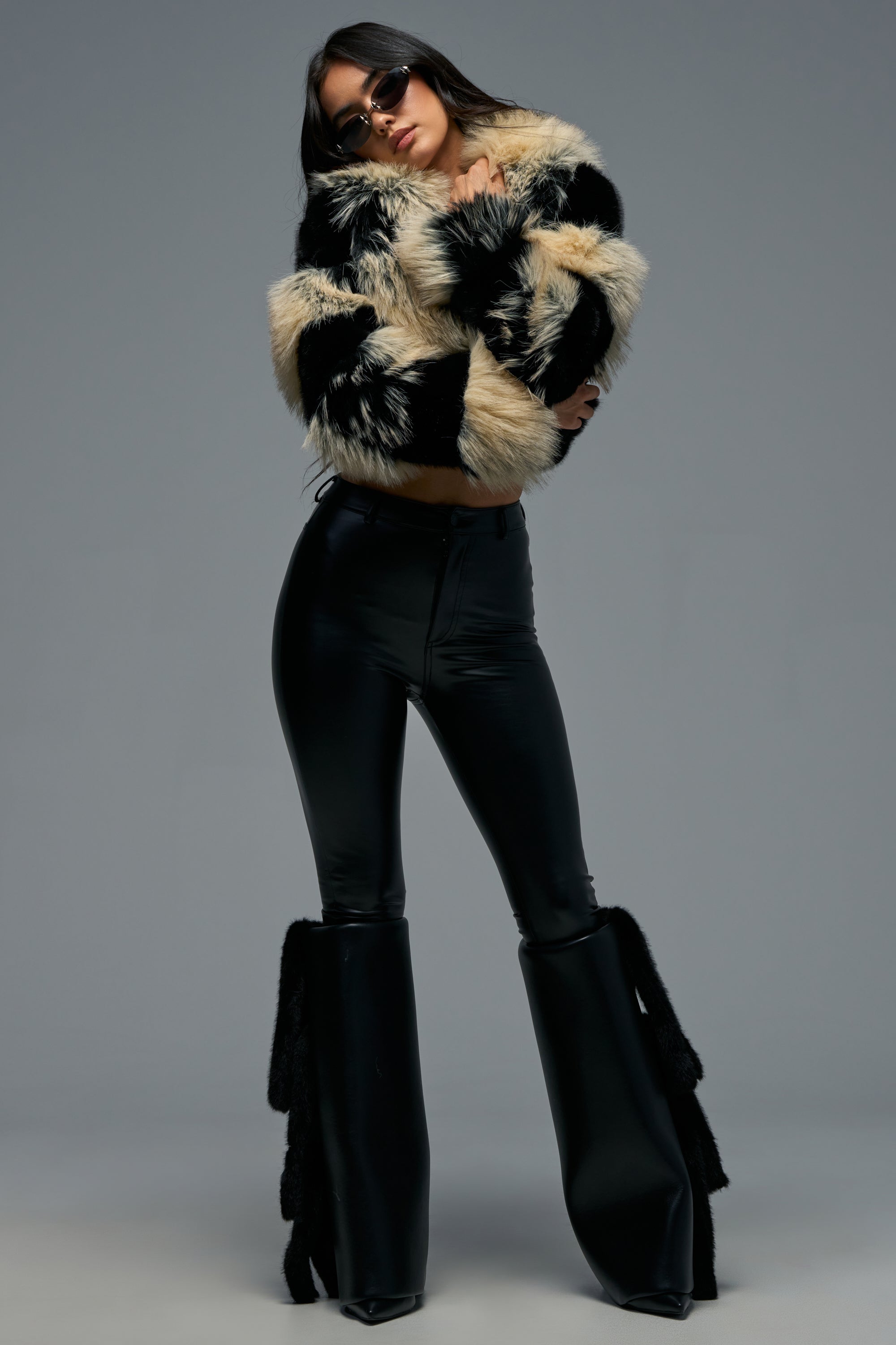 A person poses in black flared pants, the PORTIA CROPPED FAUX FUR IN BLACK BROWN jacket, and sunglasses against a plain gray background.