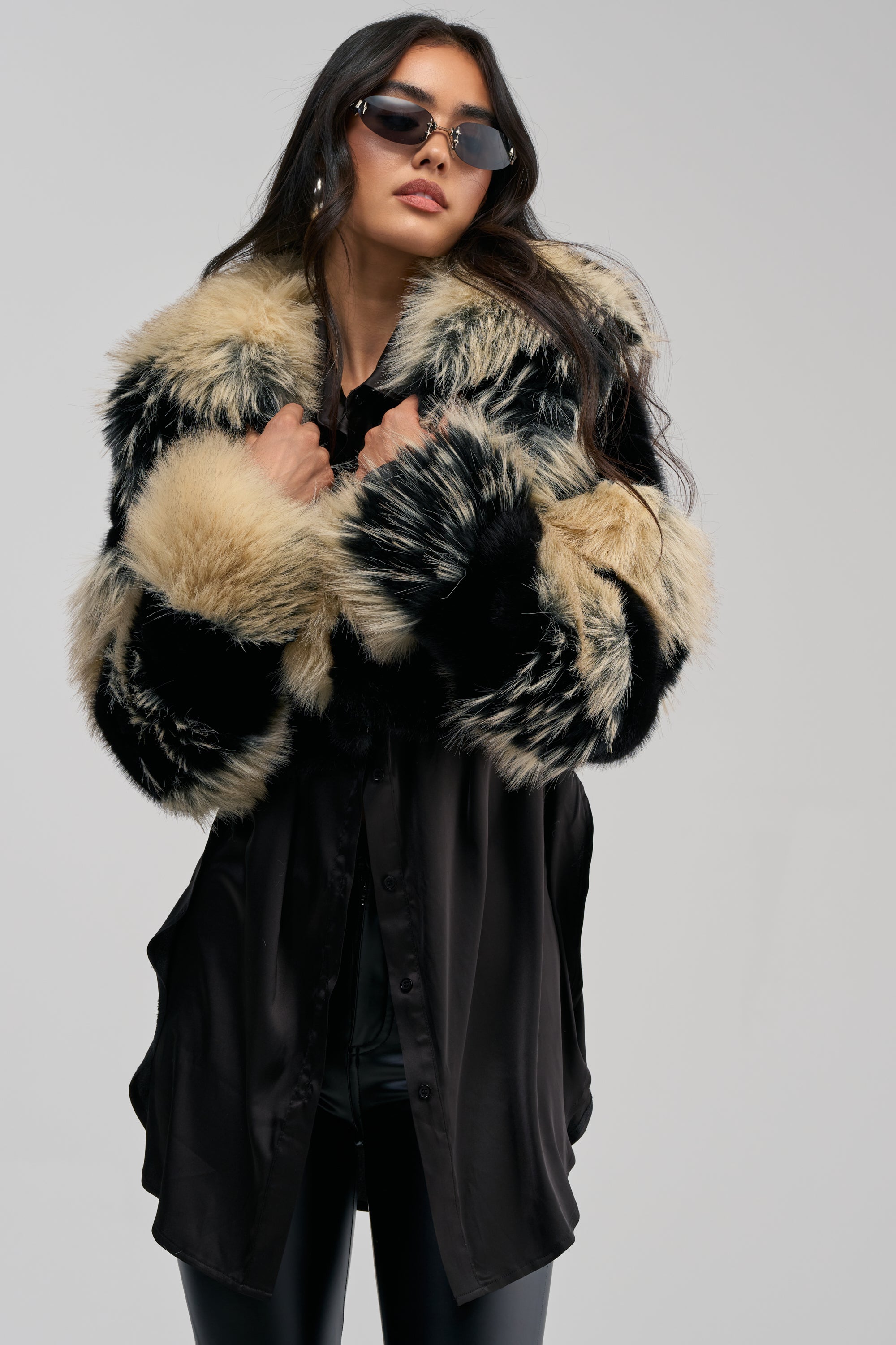 Wearing the PORTIA CROPPED FAUX FUR IN BLACK BROWN and stylish sunglasses, a woman poses confidently against a plain gray background.