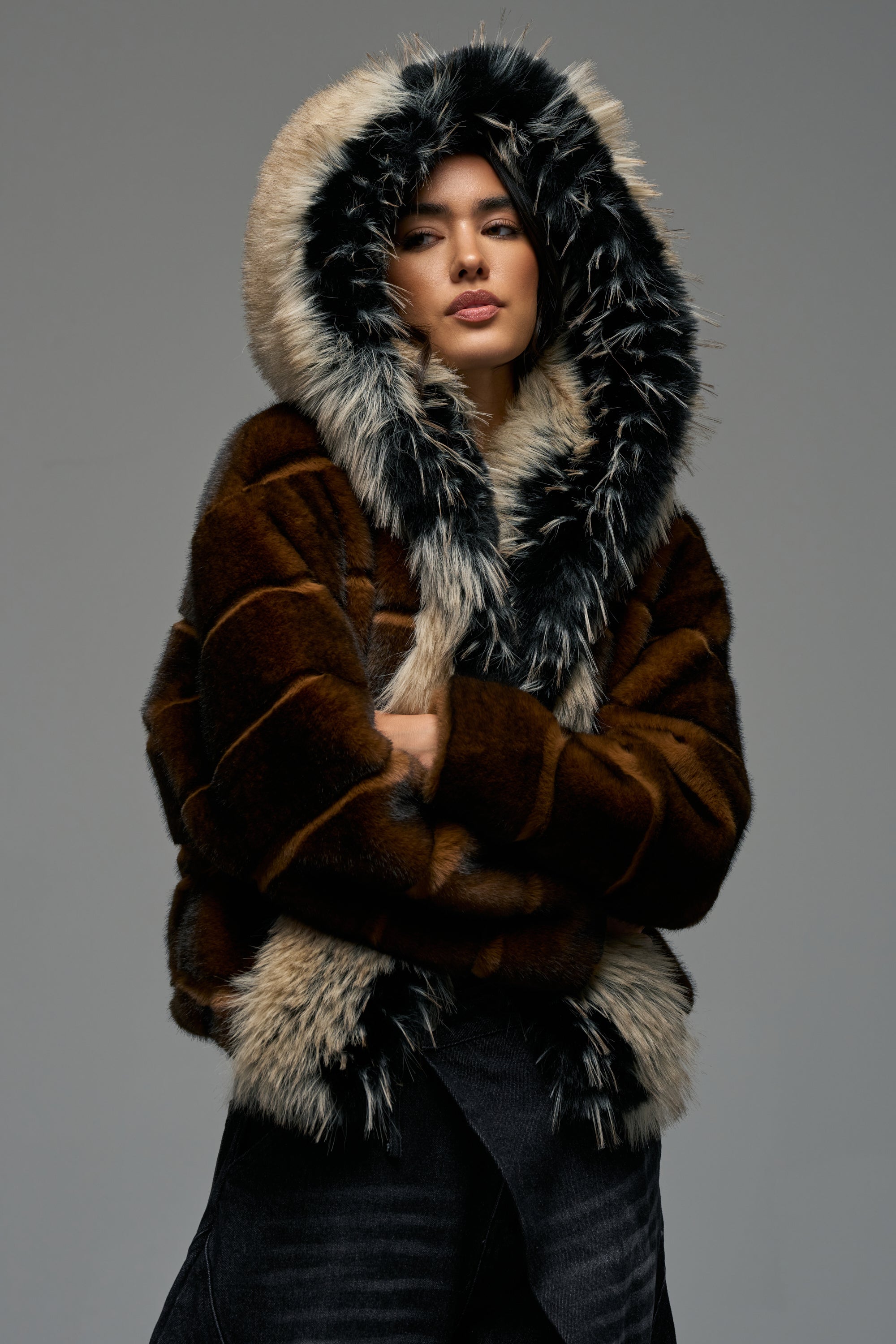 Wearing the PORTIA HOODED FAUX FUR BOMBER—a brown striped jacket styled as a fur coat with a large hood trimmed in fluffy black and white faux fur—a person stands against a plain gray background, arms crossed.