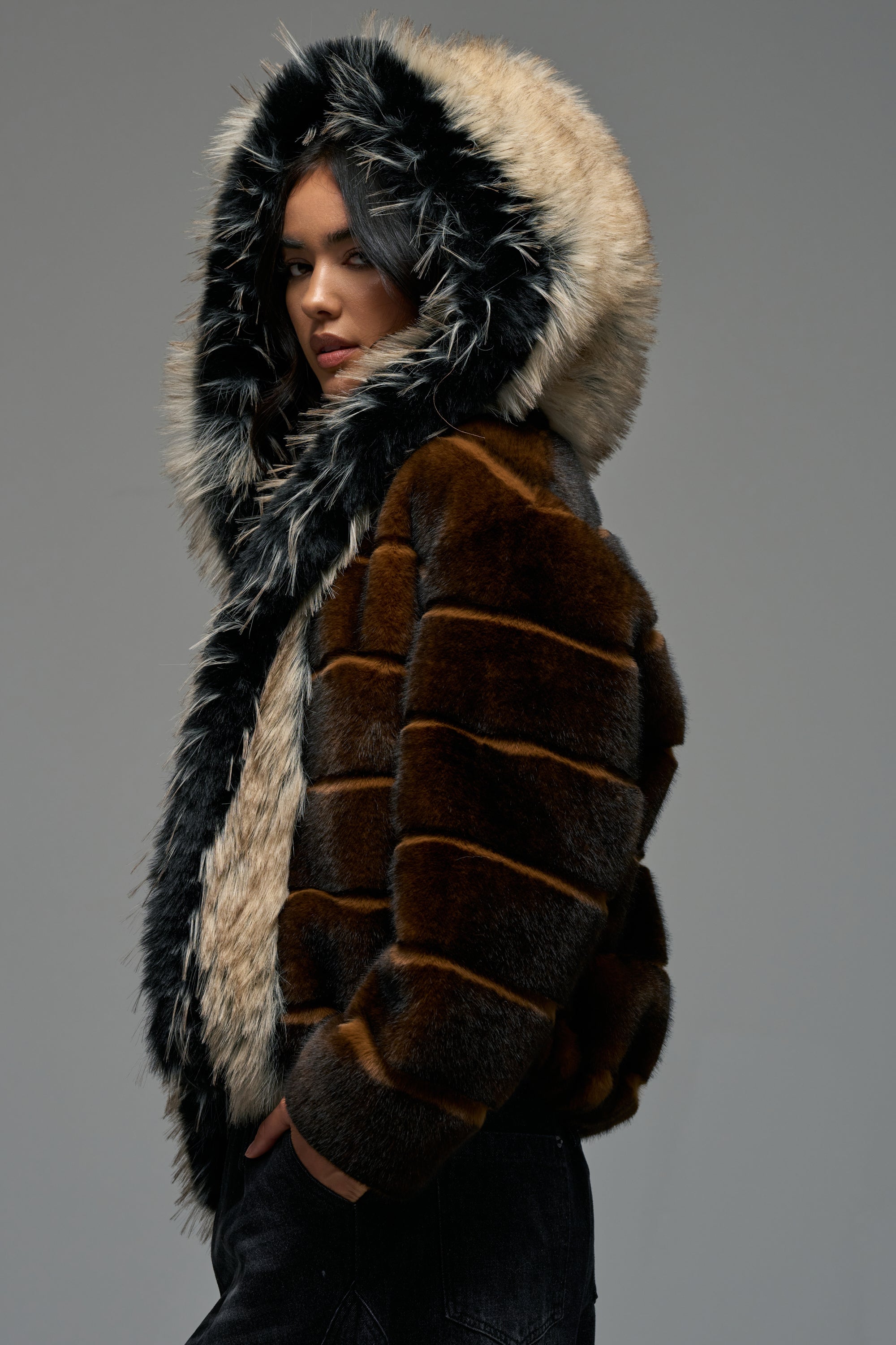 Wearing the PORTIA HOODED FAUX FUR BOMBER, a person stands against a plain gray background, looking to the side with a serious expression.