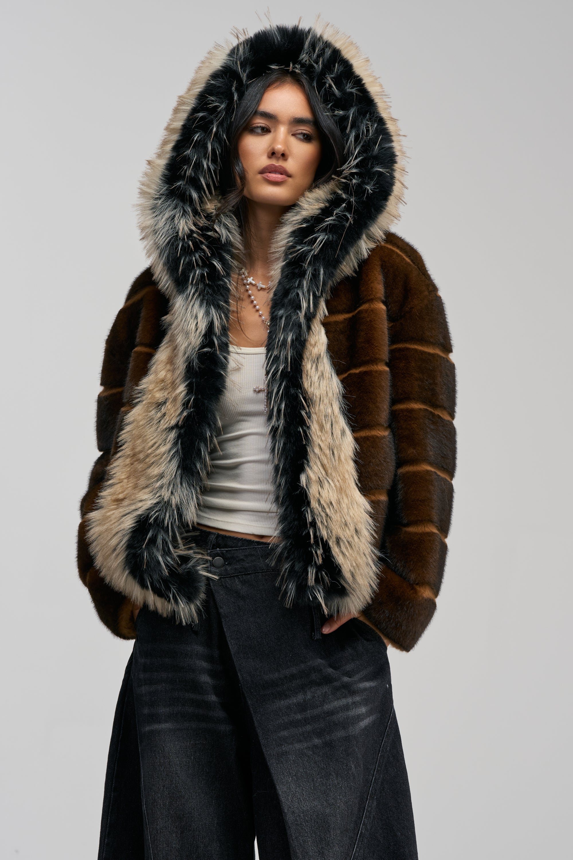 A woman wearing the PORTIA HOODED FAUX FUR BOMBER—a brown hooded jacket with black-and-white trim—along with a white tank top, dark wide-leg jeans, and a necklace stands against a plain gray background.