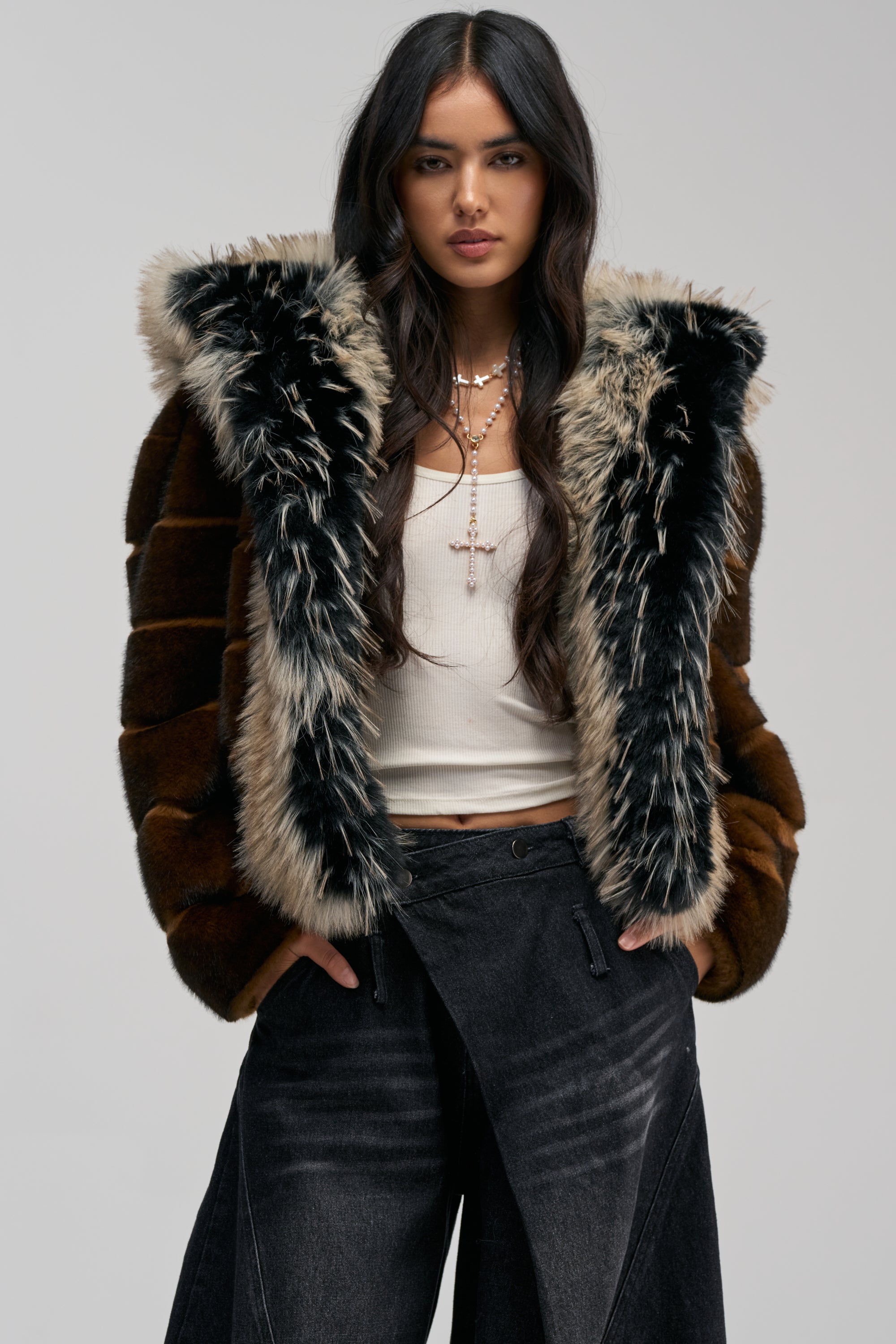 A woman with long dark hair wears the PORTIA HOODED FAUX FUR BOMBER over a white ribbed tank top, layered necklaces, and loose black pants, standing with hands in her pockets against a plain background—an effortless example of streetwear layering.