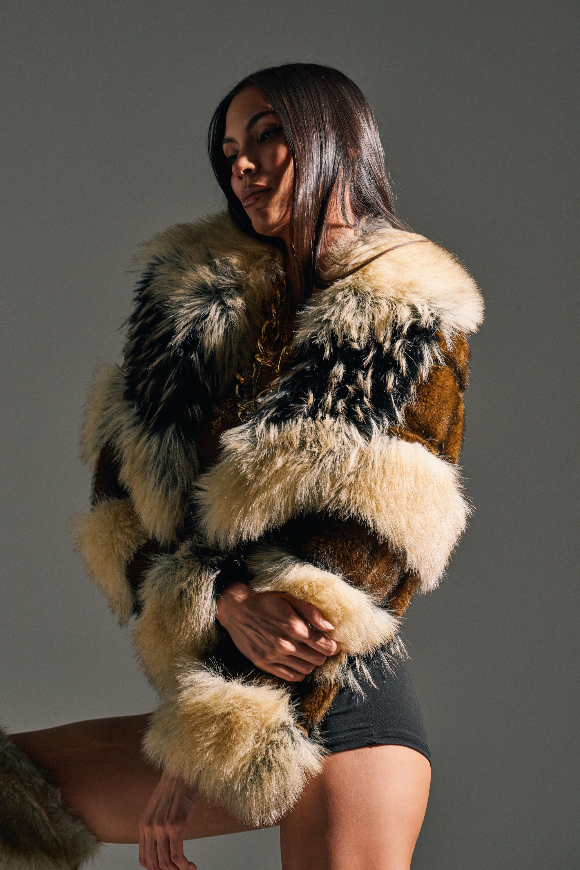 A woman with long, straight hair poses confidently against a plain gray background, wearing the PORTIA CROPPED FAUX FUR JACKET and black shorts.