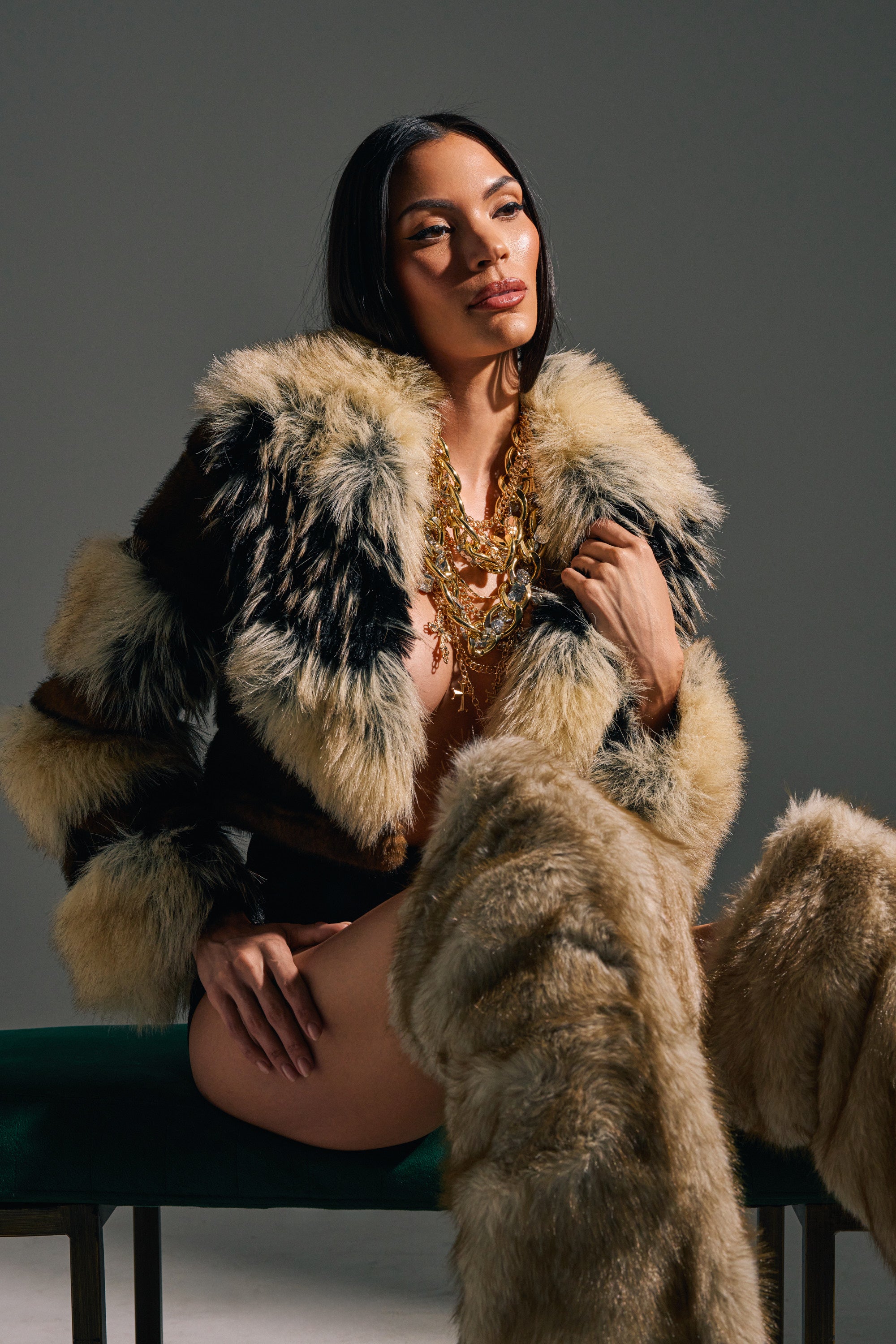 A woman with long dark hair sits on a bench wearing the PORTIA CROPPED FAUX FUR JACKET, matching fur boots, and layered gold necklaces. With legs crossed, she gazes confidently into the distance against a plain background.