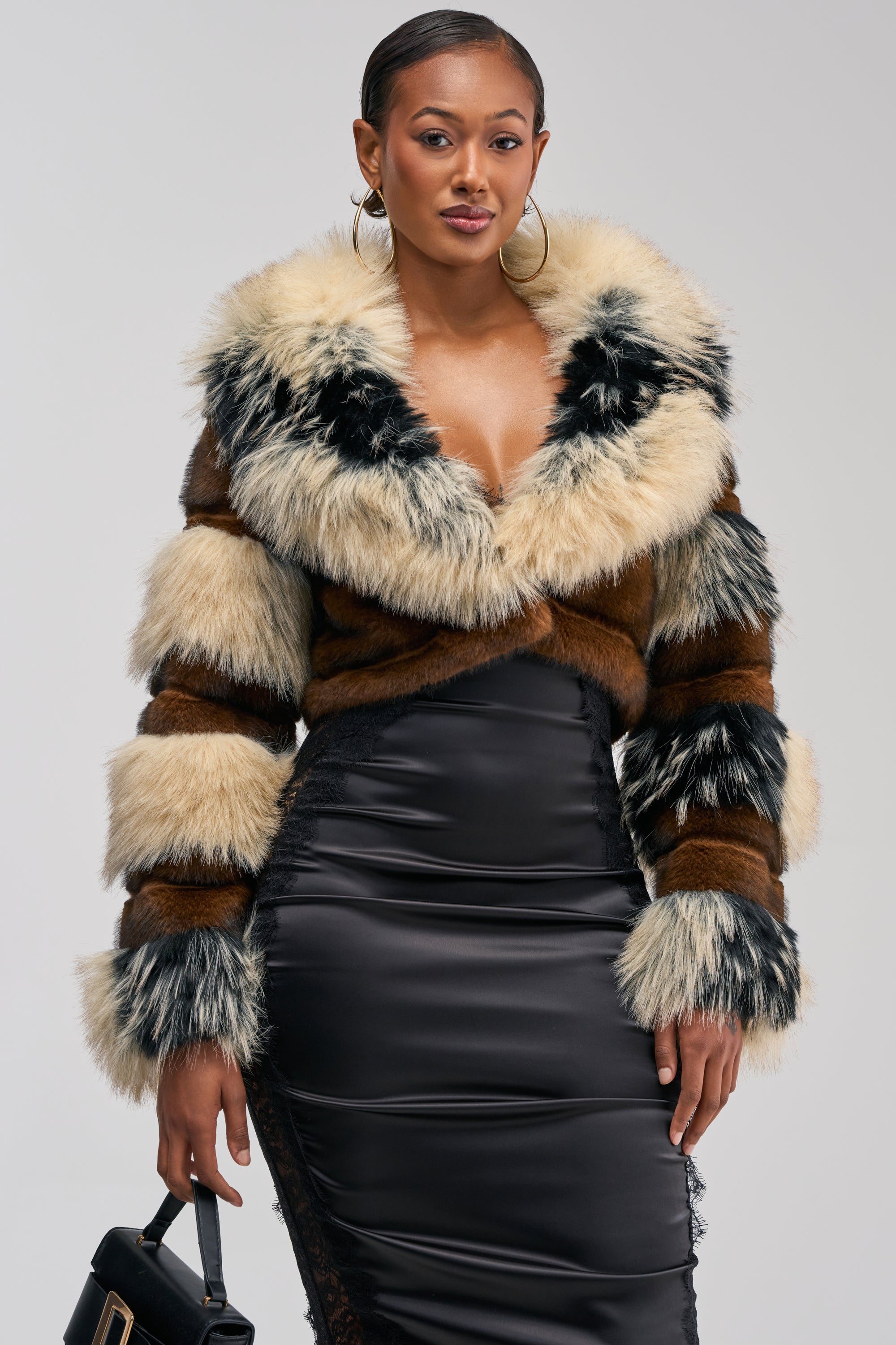 Wearing the PORTIA CROPPED FAUX FUR JACKET—a fluffy, striped faux fur piece—over a black satin dress, a woman stands confidently against a plain background holding a black handbag. The jacket’s cropped silhouette adds a modern edge to her sophisticated winter streetwear ensemble.