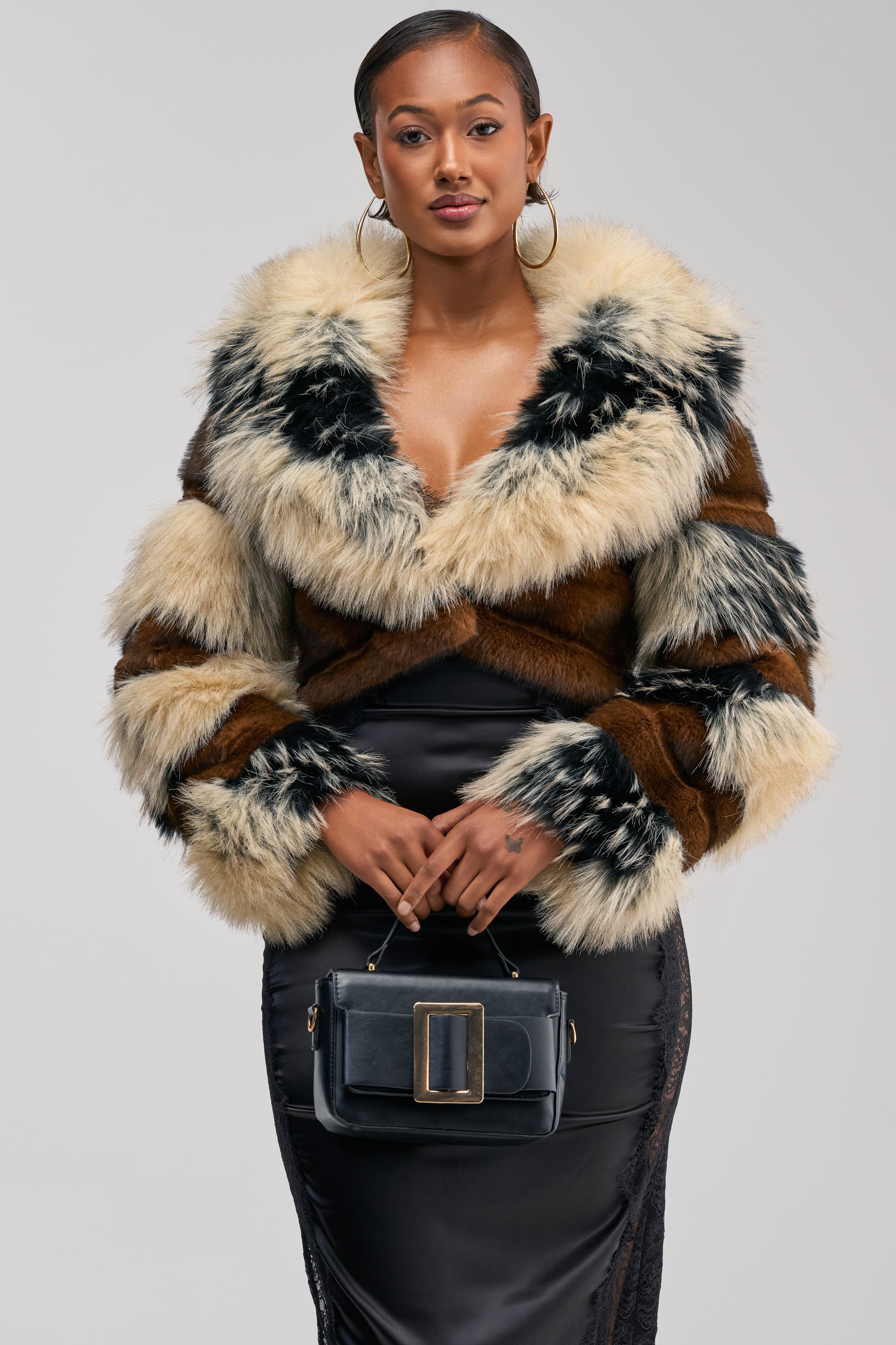 A woman showcases the PORTIA CROPPED FAUX FUR JACKET—a luxurious, multi-colored faux fur piece—layered over a black dress and paired with a statement black handbag featuring a large buckle. Her cropped silhouette brings an on-trend edge to this standout winter streetwear ensemble, all set against a neutral background.