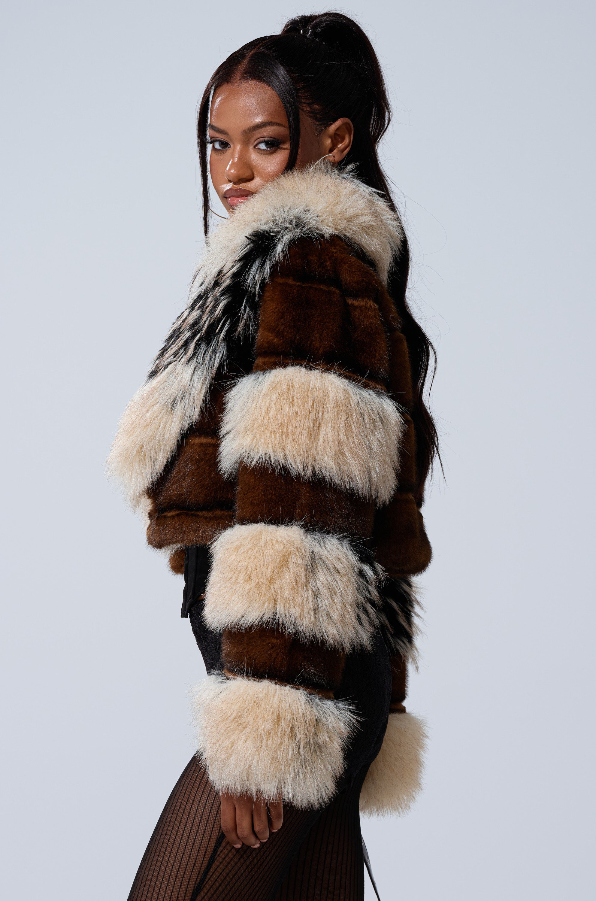 A woman with long dark hair poses sideways in the PORTIA CROPPED FAUX FUR JACKET, featuring brown, cream, and black stripes against a plain light gray background.