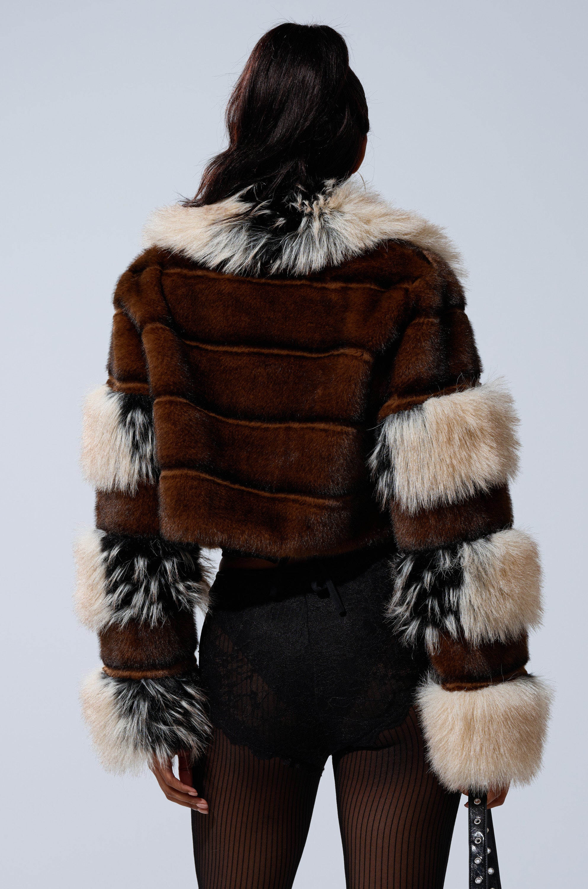 A person with long dark hair, seen from behind, wears the PORTIA CROPPED FAUX FUR JACKET featuring paneled fur and striped sleeve accents, paired with black shorts and sheer black tights.