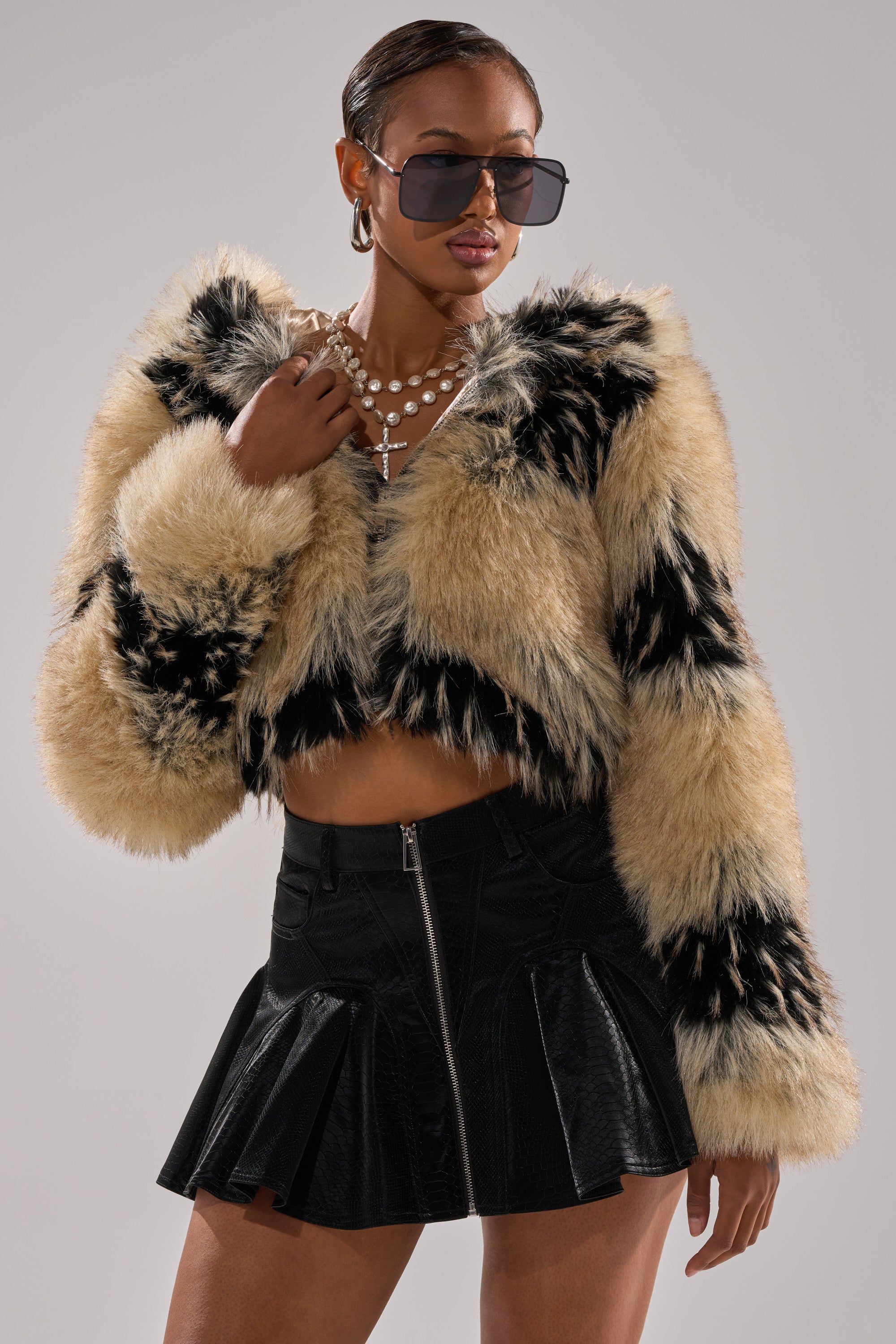 A confident woman in the ALEXIA FAUX FUR BOMBER with black and tan patterns, a black pleated mini skirt, layered necklaces, and oversized sunglasses poses against a plain background.