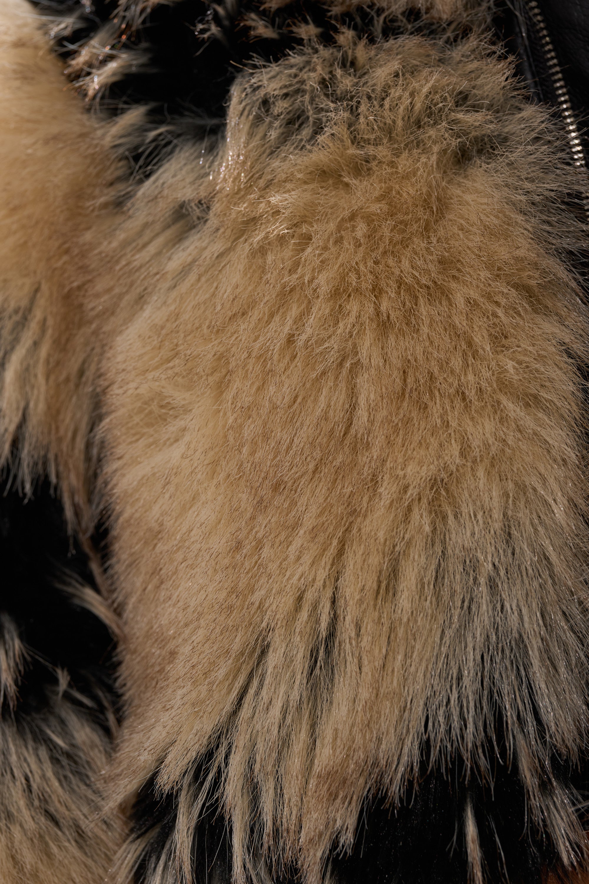 Close-up of fluffy, light brown faux fur texture on a black fabric background, likely from the ALEXIA FAUX FUR BOMBER, with visible strands and a soft look.