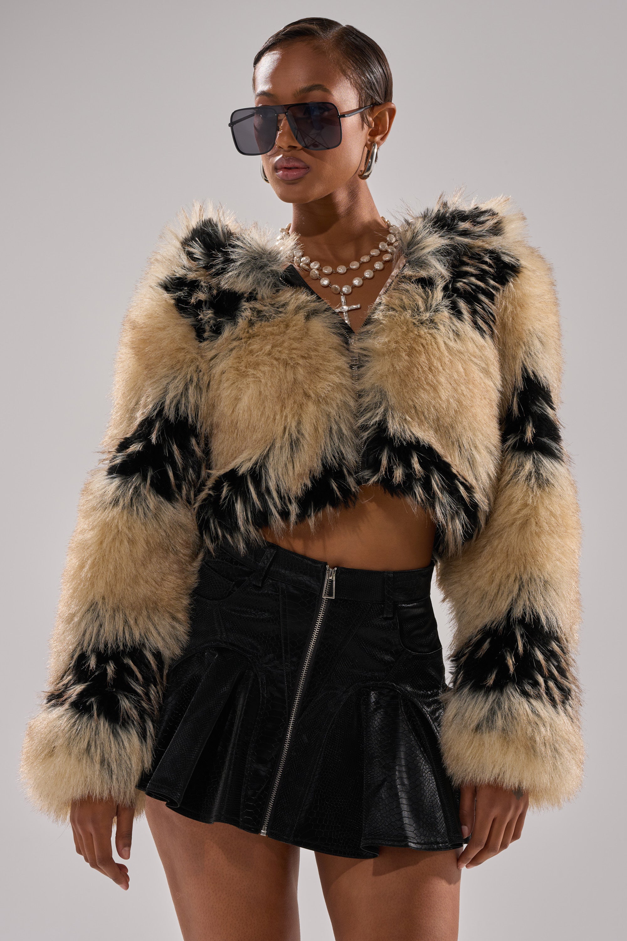 Woman wears the ALEXIA FAUX FUR BOMBER with black and tan patches, a black mini skirt with a front zipper, large sunglasses, a pearl necklace, and a cross pendant, posing against a plain background.