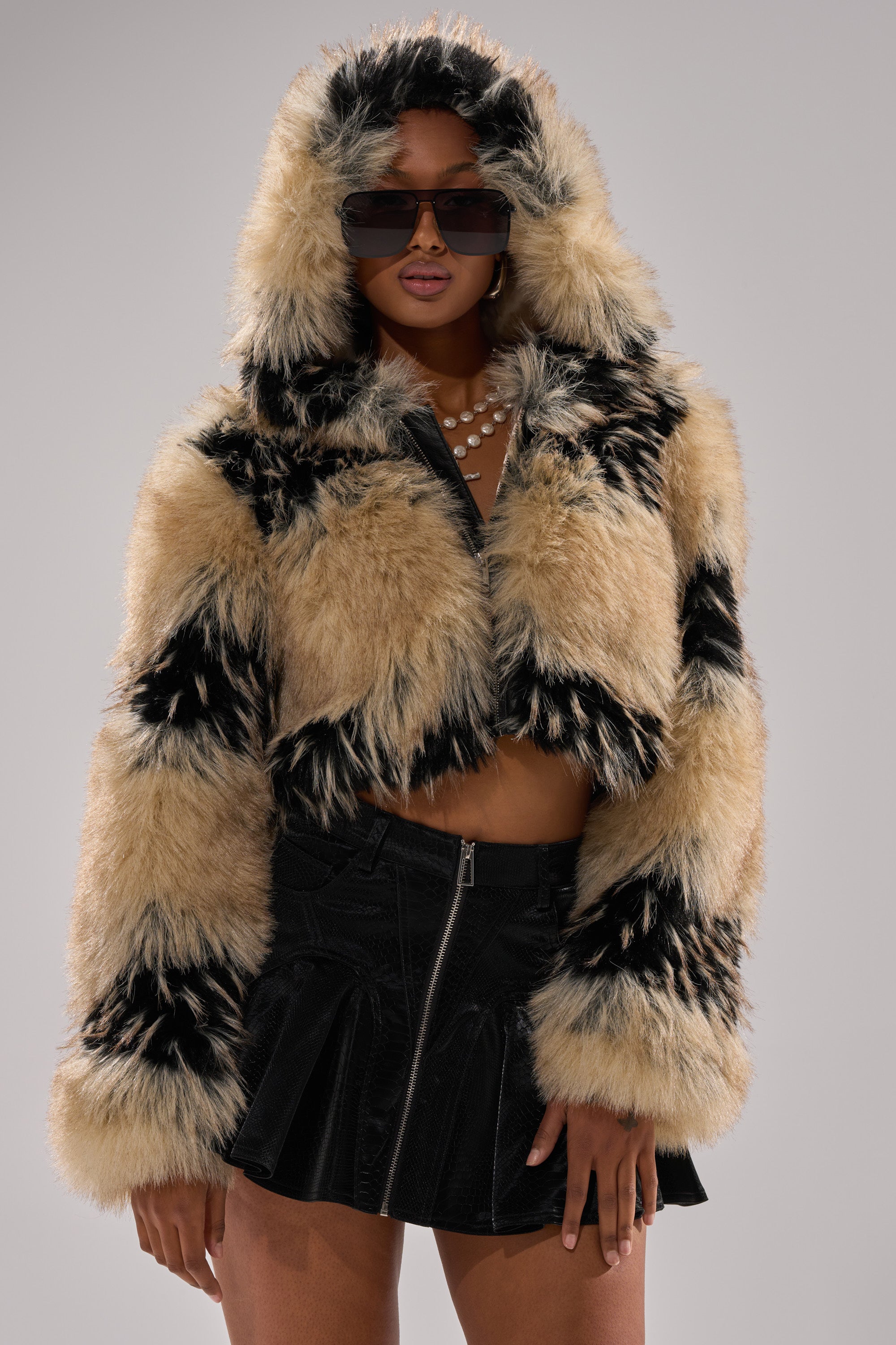 A person wearing the ALEXIA FAUX FUR BOMBER with black and tan patterns, black sunglasses, a pearl necklace, and a black pleated mini skirt stands against a plain gray background.