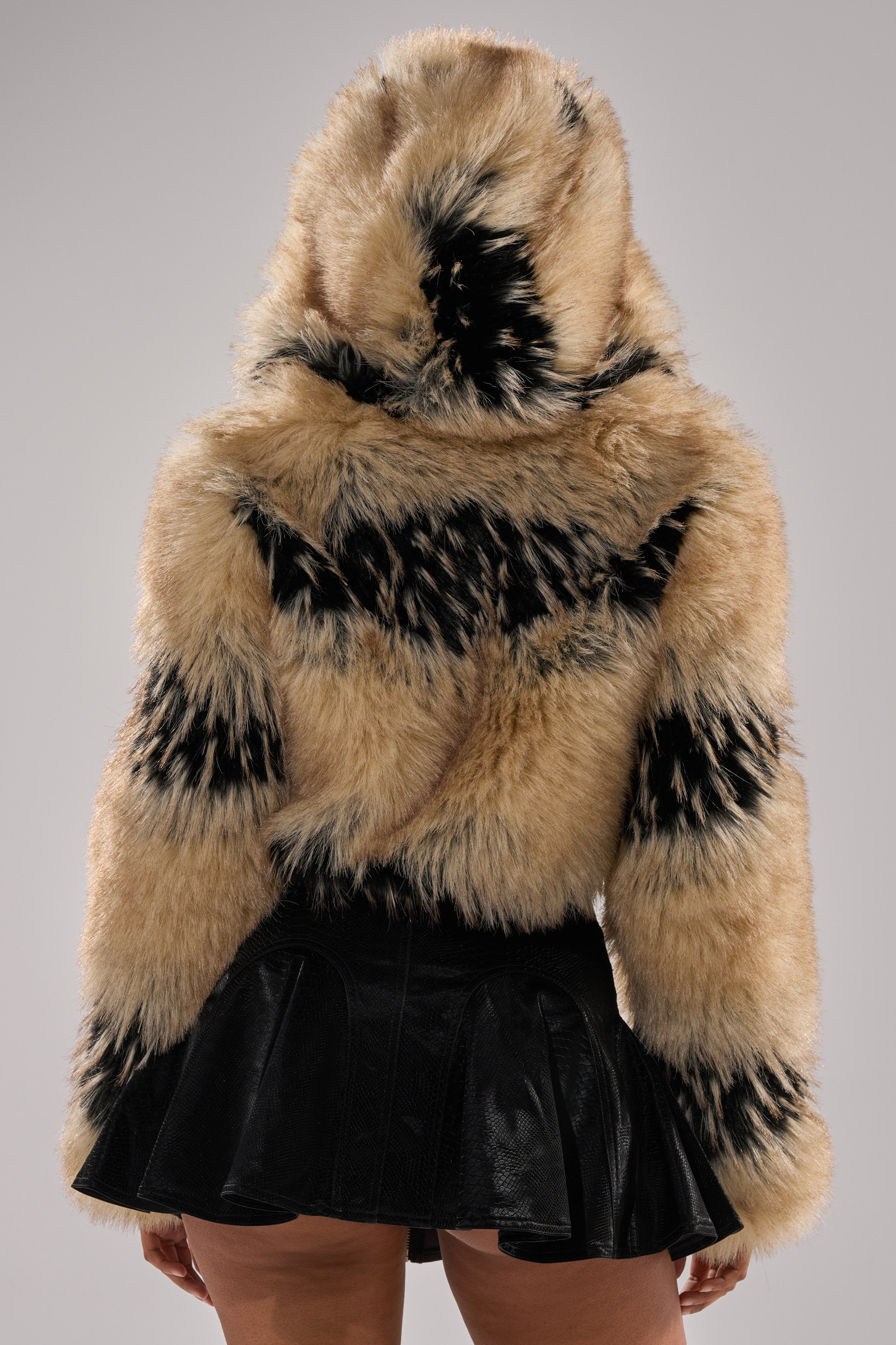 A person is seen from the back against a plain gray background wearing a tan and black ALEXIA FAUX FUR BOMBER with a hood, paired with a short black pleated skirt.