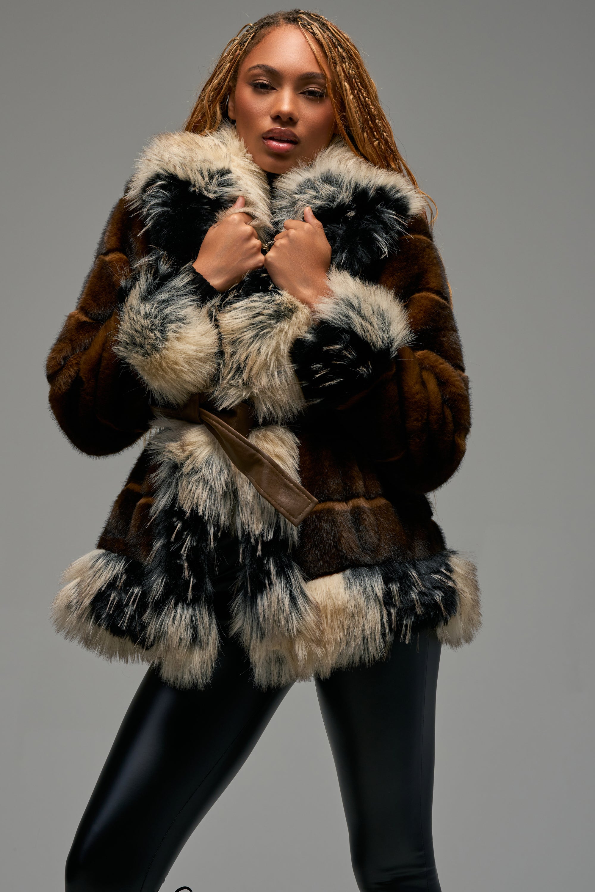 A woman with long braided hair stands against a plain grey background, wearing the KENZ FAUX FUR JACKET—featuring black, brown, and white color-block patches—and black leather pants. She holds the collar of her coat with both hands.